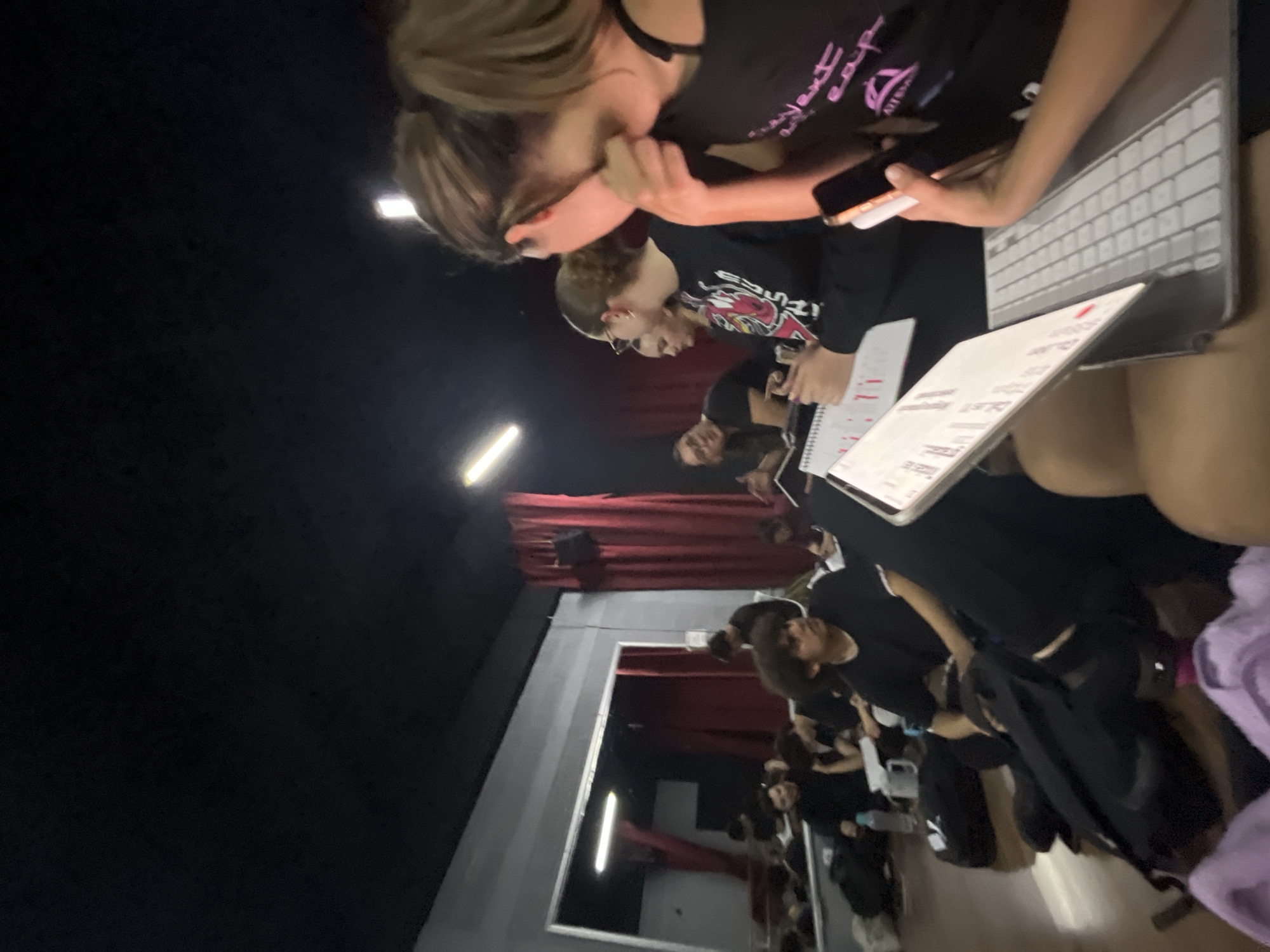 The image shows a group of people in what appears to be a dance studio or performance space in San Bernardino Tlaxcalancingo, Mexico. Red curtains are visible in the background. The people are mostly wearing black clothing. Some individuals are holding notebooks and electronic devices. The scene appears to be an observation or rehearsal.