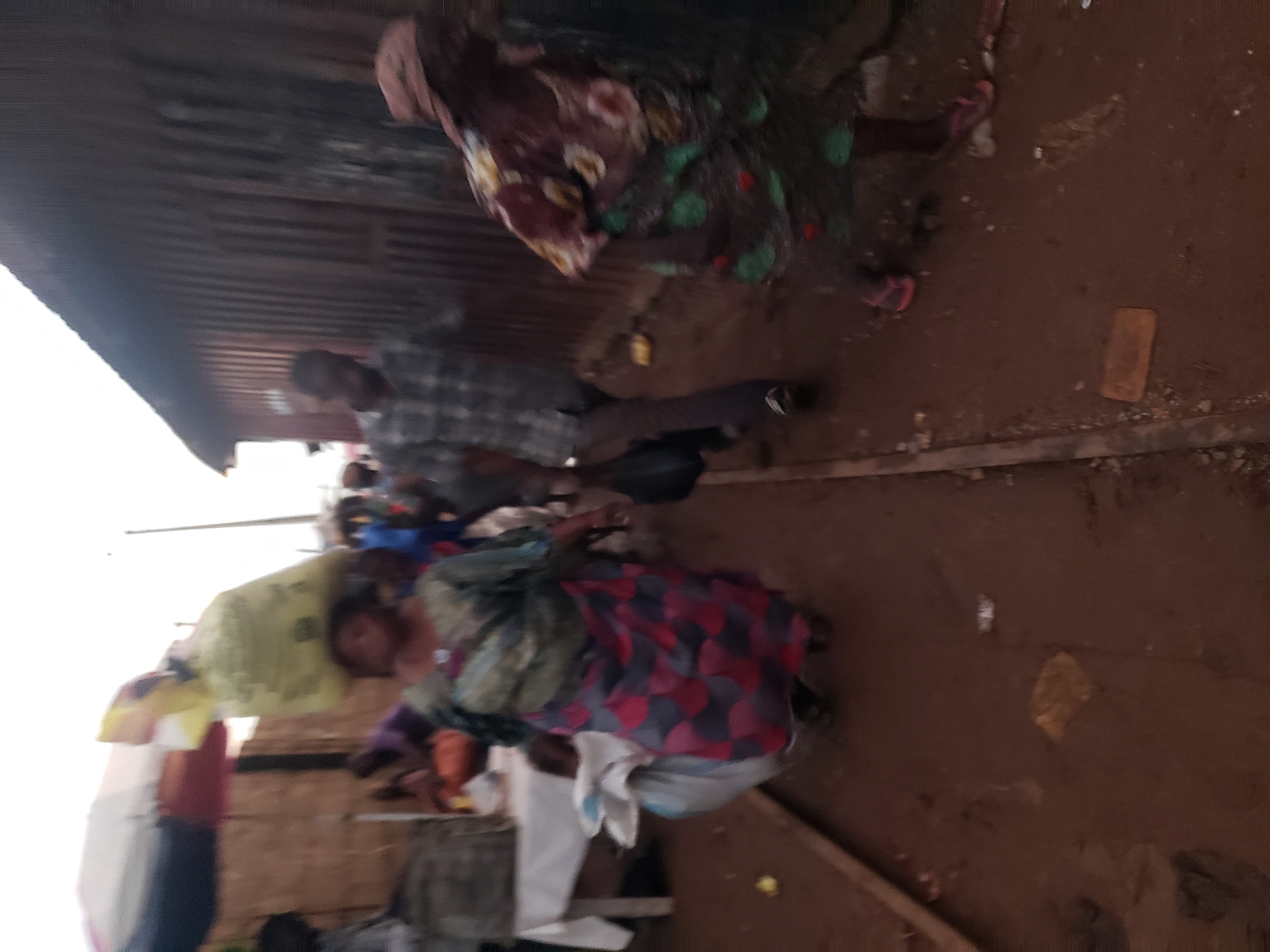 A group of people are walking on a dirt path in what appears to be a marketplace in Kakuri Gwari, Nigeria. The path is lined with makeshift stalls or structures. In the foreground, a woman with a large yellow bag balanced on her head, wearing a green and pink patterned wrap skirt and a green patterned top, walks towards the right. To her left, a man in a plaid shirt and dark pants walks away from the camera. Further to the right, another person wearing a colorful patterned dress and pink flip-flops is visible. The ground is uneven and appears muddy in some areas, with some litter scattered around. The overall lighting suggests it is daytime.