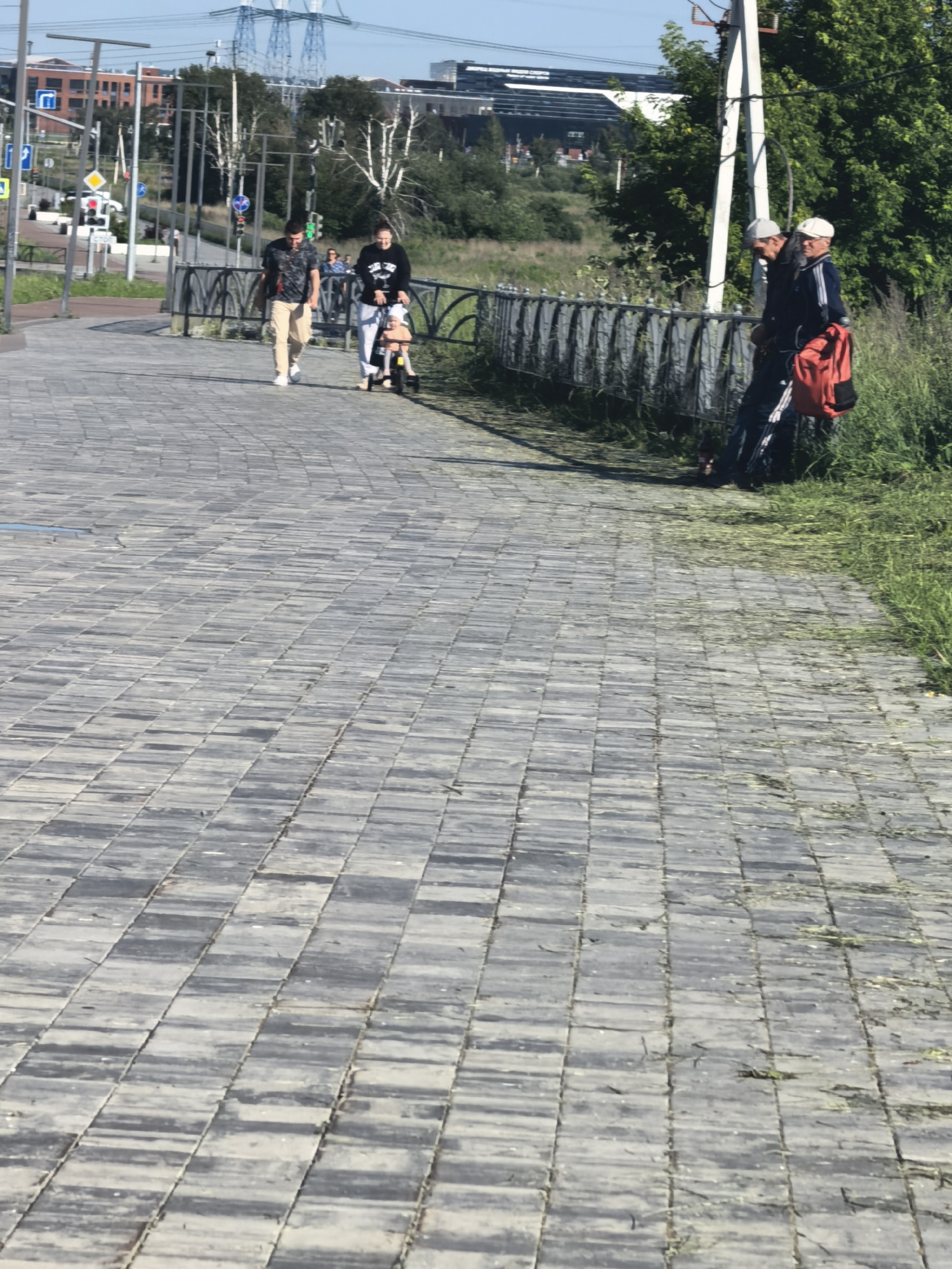 The image shows a paved walkway in a city, with a grassy area and a fence on the right side. In the foreground, the walkway is made of rectangular grey pavers laid in a basketweave pattern. On the walkway, a man wearing a dark patterned shirt and light-colored pants walks towards the viewer, carrying a bag. Behind him, a woman in a black sweatshirt pushes a small stroller with a child. Further back, two men in athletic wear, one with a white cap and the other with a white baseball cap, are walking towards the right. One of the men is carrying a bright orange backpack. The background shows trees, buildings, and electrical towers. The overall scene depicts a sunny day in an urban park or promenade.