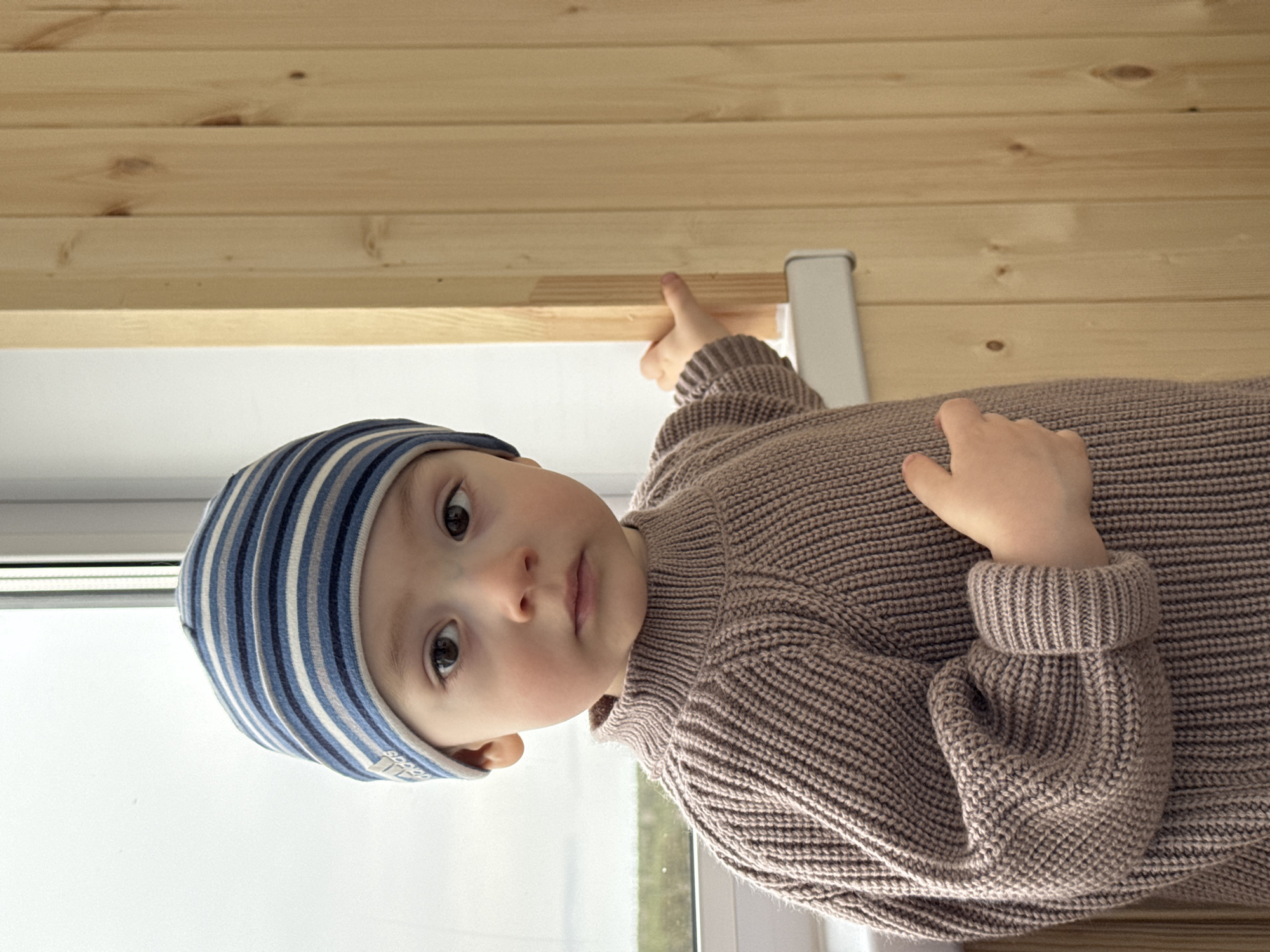 A young child, wearing a blue and white striped hat and a brown knit sweater, is positioned in front of a window and wooden wall or ceiling. The child is looking directly at the camera with a serious expression. The location is not identifiable as Izhevsk, Russia from the given image. The child's right hand is raised, touching the wooden structure above, while the left hand rests on their chest. The background consists of the window and the light-colored wood, which appear to be part of an interior structure.