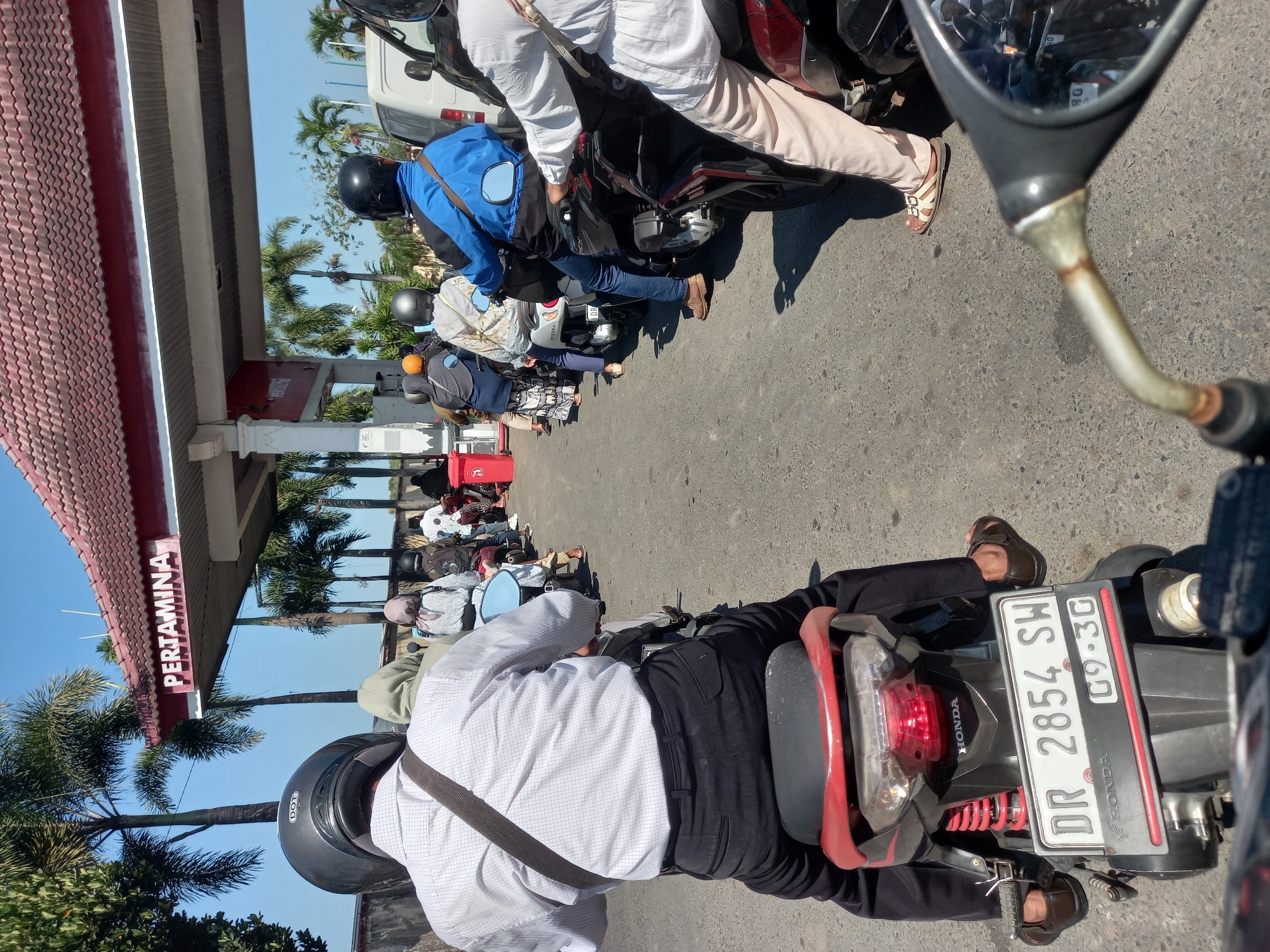 In Mataram, Indonesia, on a sunny day, a queue of motorcyclists waits at a Pertamina gas station. The foreground shows the back of a motorcycle with a red and black body, a license plate reading "DR 2854 SH 09.30", and a rider wearing black pants and brown sandals. Following behind are several other motorcyclists, some with passengers, on various models of scooters and motorcycles. They are all heading towards the covered area of the gas station where the pumps are located. Palm trees line the perimeter of the station, and a white van is visible further down the line. The scene is typical of a busy afternoon at a gas station in the city.