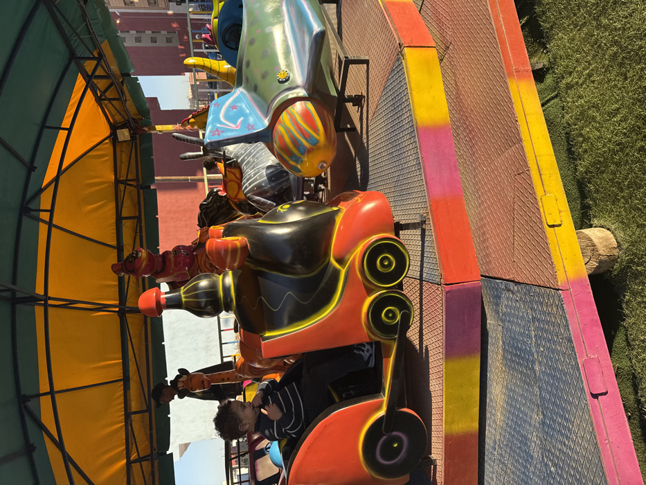 This outdoor, brightly lit image captures a segment of a children's amusement park ride, likely located in Ait Ouallal, Morocco. The scene features a colorful track with several ride vehicles and a young child as the primary subject.
In the foreground, a young child with dark, curly hair, wearing a striped long-sleeved shirt, sits in an orange and black ride vehicle with yellow accents. This vehicle has a futuristic or robotic design, featuring prominent circular light-like elements at the front. The child is looking to the left, with one hand near their mouth, suggesting a moment of observation or anticipation. Further along the track, other ride vehicles are visible, including a green and white one with a striped snout, possibly resembling a fish or alien creature, and additional animal-shaped vehicles (like a giraffe and a horse) under a canopy.
The ride track consists of textured metal sections, interspersed with vibrant, painted barriers in shades of yellow, pink, and orange. To the left, a large, arched canopy or awning, with a bright yellow fabric top and a green metal frame, provides shade over part of the ride. On the right side of the track, a patch of artificial green grass is visible. In the background, under a clear blue sky, brick-colored buildings can be seen.
The activity depicted is likely children enjoying or waiting for a ride at an amusement park or fair. The bright sunlight and distinct shadows indicate it is daytime. No discernible text is visible within the image.