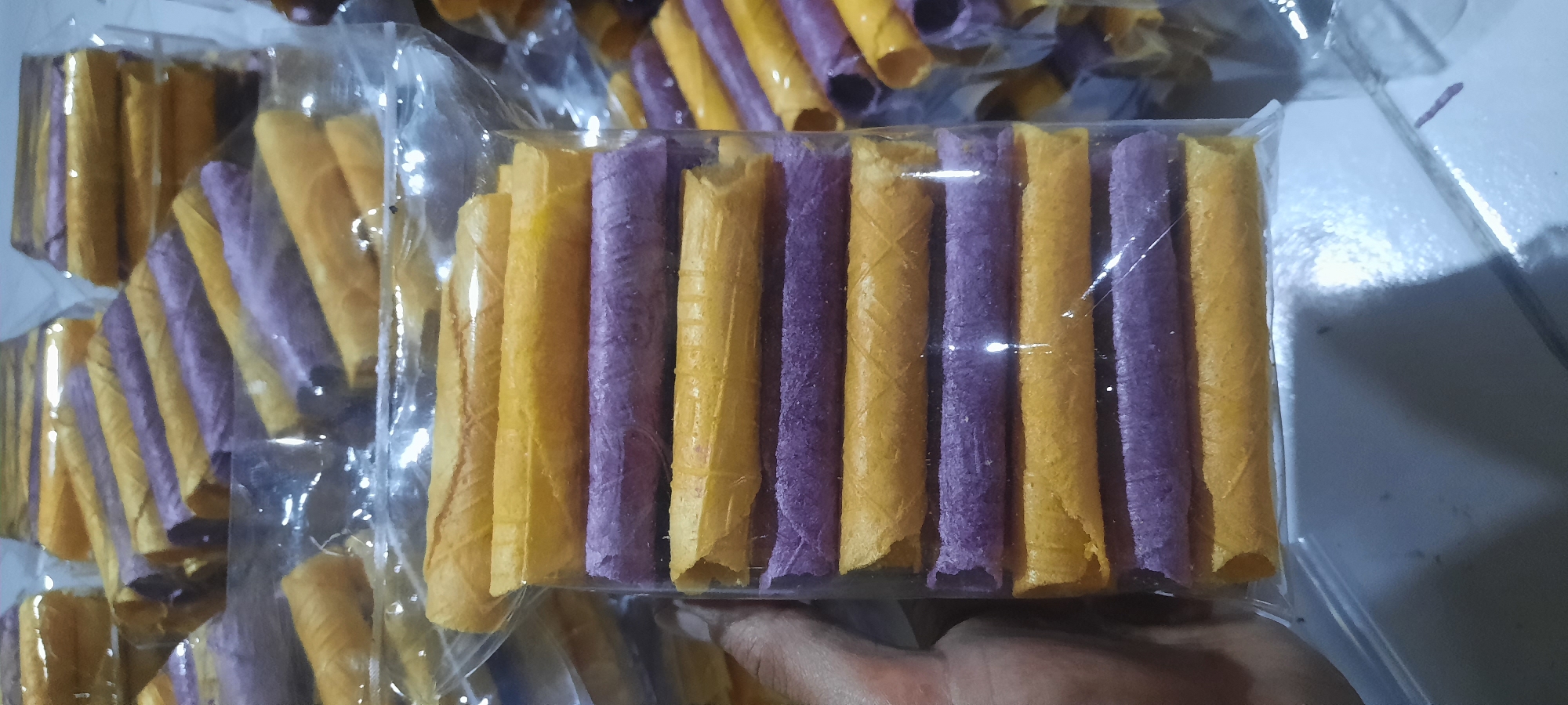 The image shows a close-up view of several clear plastic packages containing rolled wafer cookies. The cookies are arranged in stacks within the packages, alternating between yellow and purple colors. A person's left hand is visible on the left side of the frame, touching the package. The background is out of focus but appears to be a white or light-colored surface. The scene is indoors, likely a retail or home setting. There are no specific landmarks or environmental cues to indicate a precise location within Ploso, Indonesia.