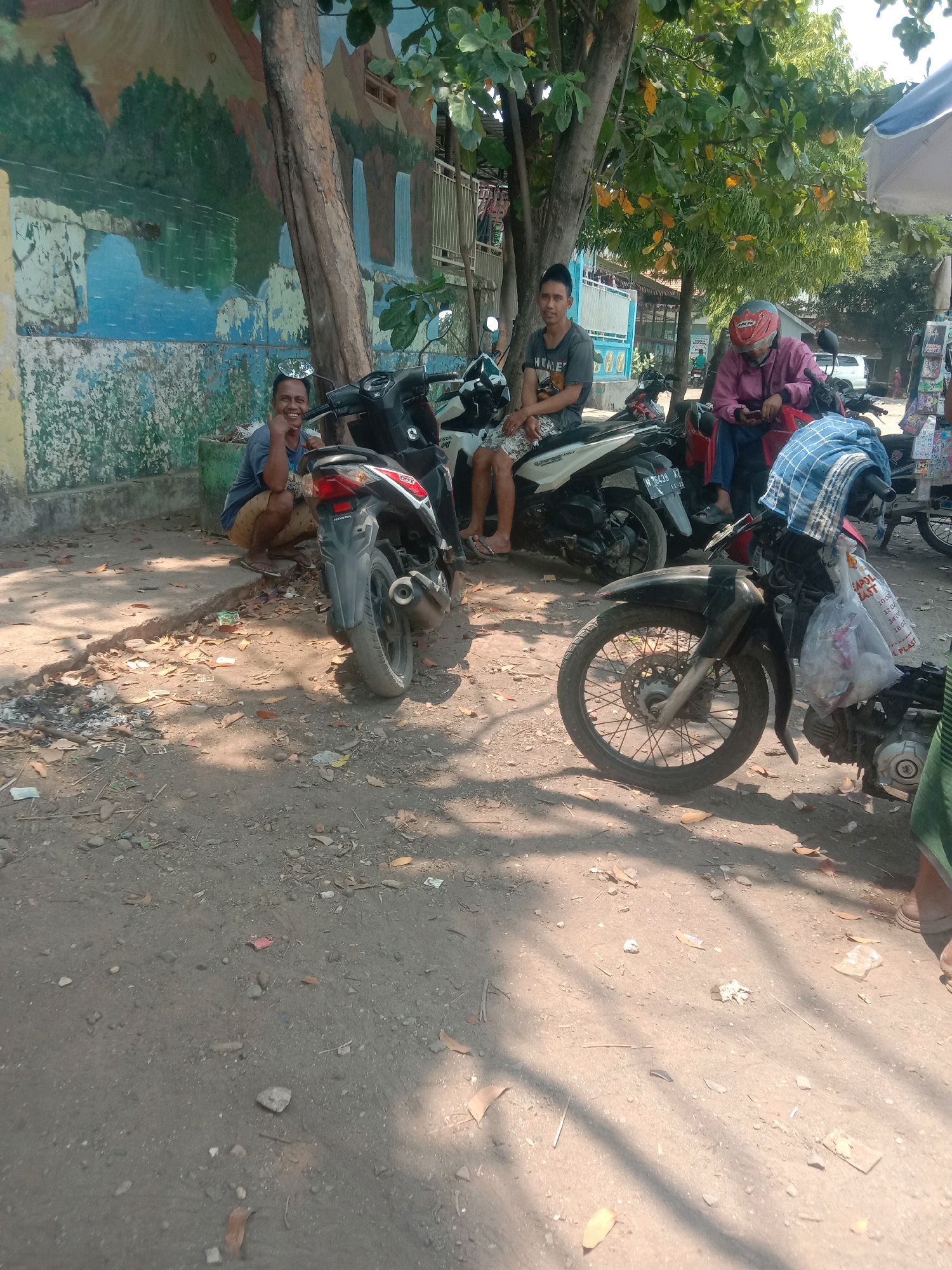 In Ngempit, Indonesia, two men are seen near motorcycles parked on a dirt road. One man, wearing a blue t-shirt and brown shorts, is crouched down and smiling, with a reflective disc on his head. The other man, wearing a grey t-shirt and patterned shorts, sits on a white motorcycle. A third person, wearing a pink jacket and red helmet, is seated on a red motorcycle, looking at their phone. In the background, a mural adorns a wall, depicting a landscape with trees and waterfalls. The area is cluttered with leaves and debris.