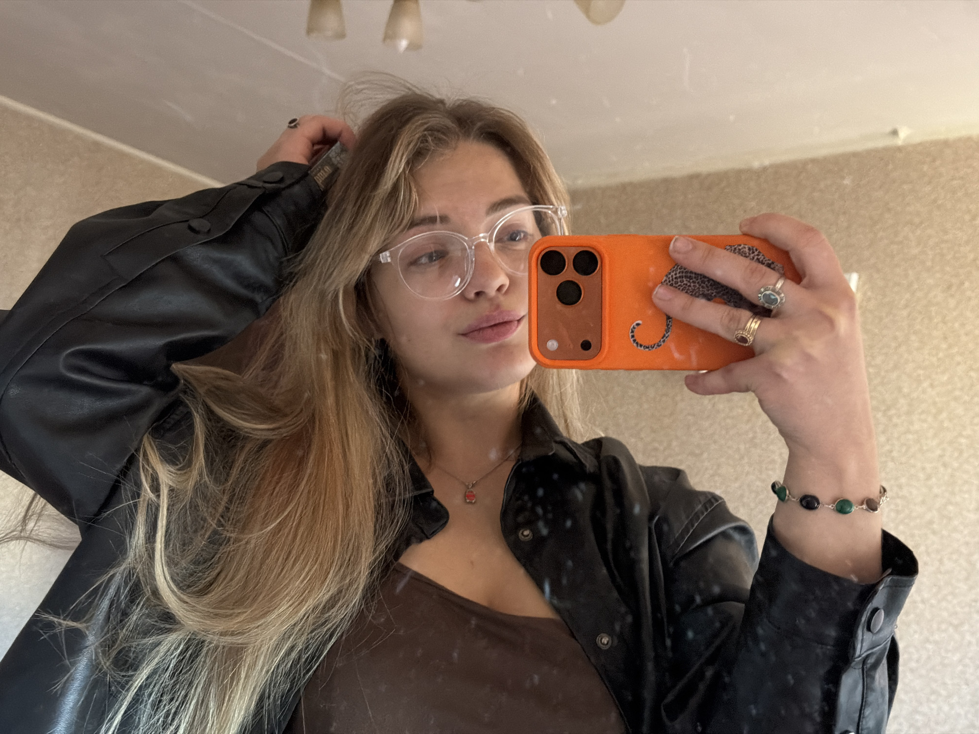 A young woman with blonde hair and clear-framed glasses is taking a selfie in what appears to be an indoor setting with light-colored walls. She is wearing a brown top and a black, button-down jacket. Her left hand is touching her hair, while her right hand holds an orange smartphone with a leopard print design on the case. Jewelry is visible, including rings on her fingers, a necklace, and a bracelet. The location is difficult to determine from the image, and without additional context, the association with Naberezhnye Chelny, Russia, cannot be substantiated.