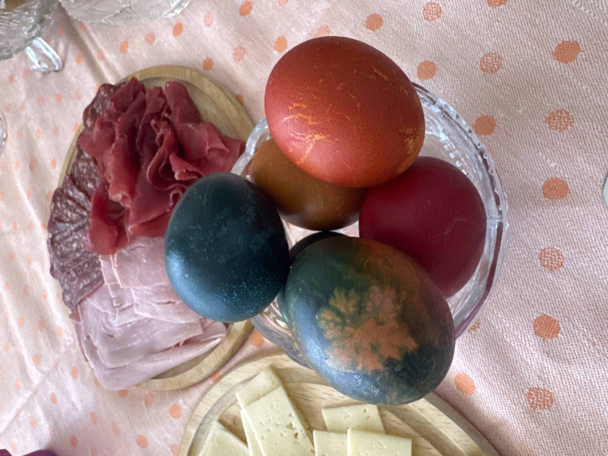 A top-down shot captures a section of a table, likely indoors, set with various food items. The tablecloth features a light pink background with an orange polka dot pattern. A clear glass bowl is partially visible, filled with five colored eggs. These eggs exhibit shades of red, brown, and dark green, with one egg displaying a mottled pattern of green and brown. Adjacent to the bowl, a circular wooden board holds arranged sliced meats, including salami, ham, and prosciutto. Another wooden board presents a selection of sliced cheese. This setting suggests a festive meal or gathering, possibly an Easter celebration given the presence of colored eggs. While specific details point towards the region, I do not have enough information to verify that it is Sofia, Bulgaria.
