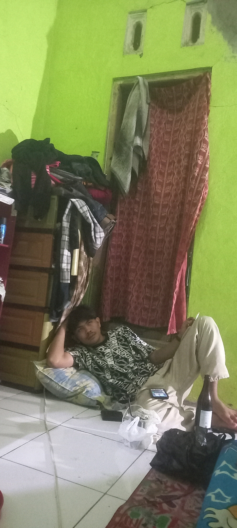 The image depicts an indoor scene within a room in Argasoka, Indonesia. A young man is seen reclining on a pillow on the floor, with his left arm propped behind his head and his right arm resting on his lap. He is wearing a patterned short-sleeved shirt and light-colored pants. In front of him, on the floor, is a smartphone, a plastic bag, and a dark bottle. Behind the man, to his left, is a tall, multi-tiered drawer unit overloaded with clothing. To his right and behind him, there is a doorway covered by a patterned red curtain, and above the doorway, two small rectangular ventilation openings are visible in the lime green wall. The floor is tiled.