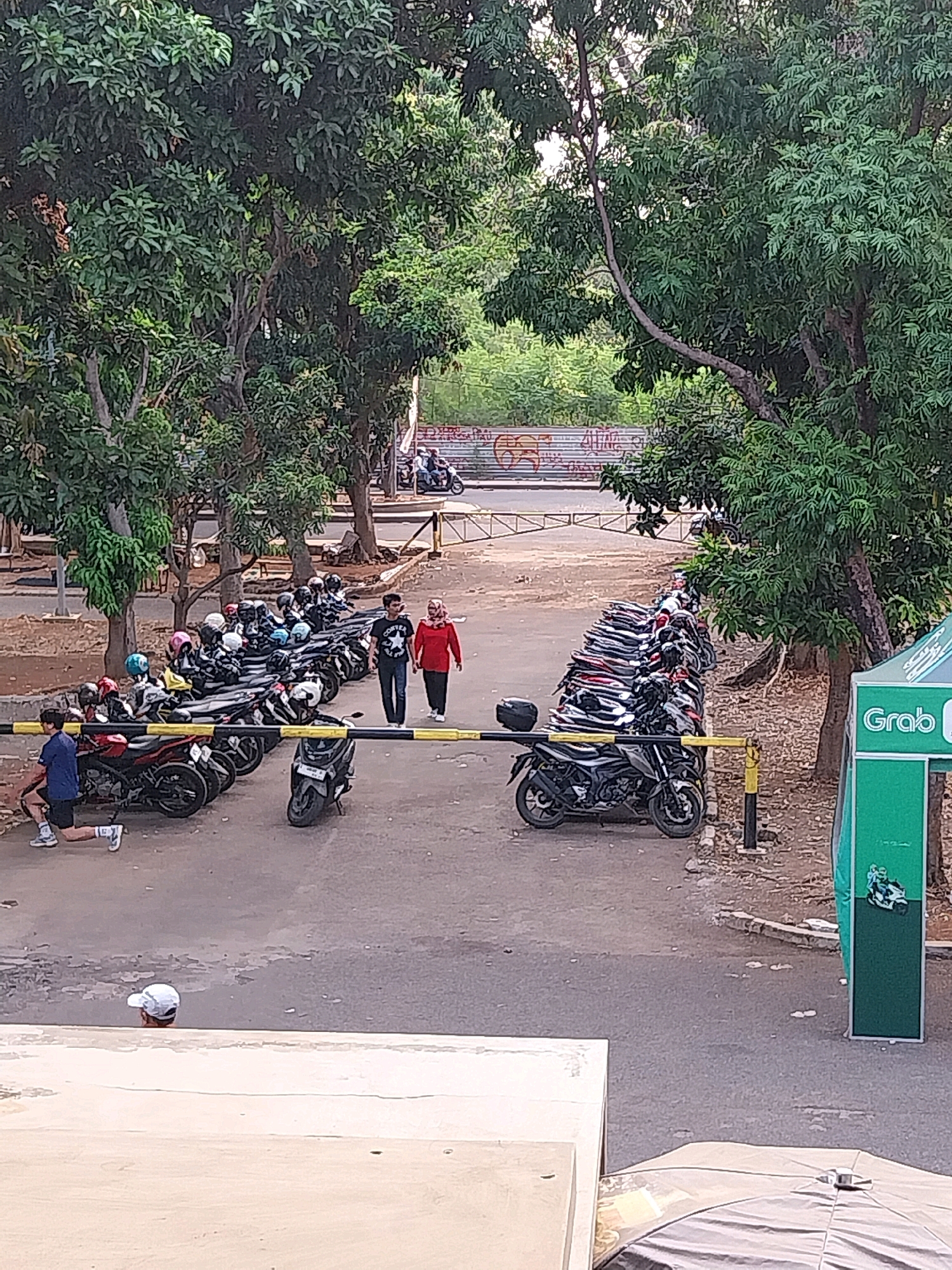 This image depicts an outdoor scene with a row of parked motorcycles under trees. In the foreground, a person is performing a lunge exercise on the asphalt. Further back, two individuals are walking side-by-side on a paved area. To the right, a green structure with the Grab logo is visible. The background shows more trees and a fence with graffiti. The overall environment suggests a parking area or a public space within a city. The presence of the Grab logo indicates a connection to ride-sharing services, which are prevalent in Indonesian cities like Cirebon. The scene appears to be captured during daylight hours.
