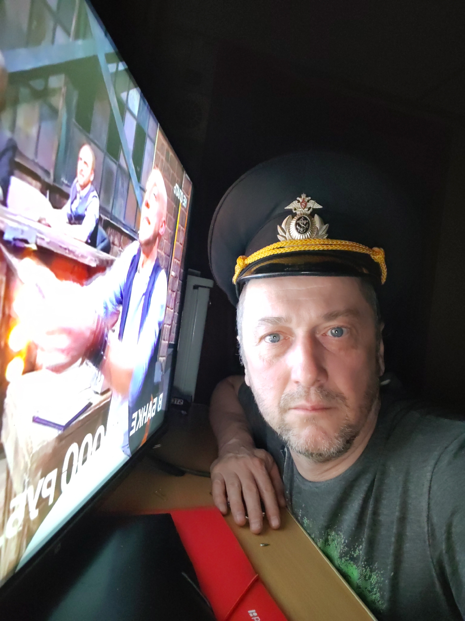 A man is shown in a selfie indoors, next to a television screen. The man is wearing a dark peaked cap with a metallic insignia and gold rope trim. He has a beard and light-colored eyes. The television screen shows a scene with two people. One person is looking up with an expression of surprise, while the other person is partially visible on the left. Text is visible on the television screen, written in a script that appears to be Cyrillic. The man is leaning on a wooden surface next to the television, with a red object visible below. The environment is dimly lit. Although the city of Ermolino, Russia is mentioned, there is no visual evidence in the image to confirm the location.
