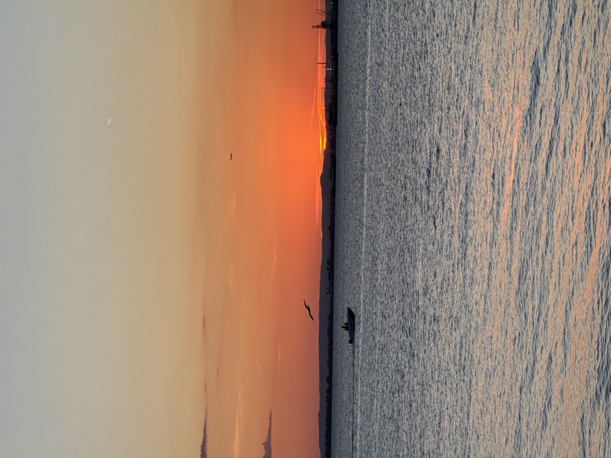 The image depicts a serene seascape at sunset in Burgas, Bulgaria. The sky is a gradient of soft blues and oranges, transitioning into a fiery red on the horizon where the sun is setting. Silhouetted against this vibrant sky are distant hills, a city skyline with industrial cranes, and a faint airplane contrail. The sea is calm, with gentle ripples reflecting the warm hues of the sunset. In the foreground, a small boat with two figures is navigating the water, leaving a subtle wake. A seagull is captured mid-flight, adding a dynamic element to the scene. The overall atmosphere is tranquil and picturesque, characteristic of a coastal sunset in Burgas.
