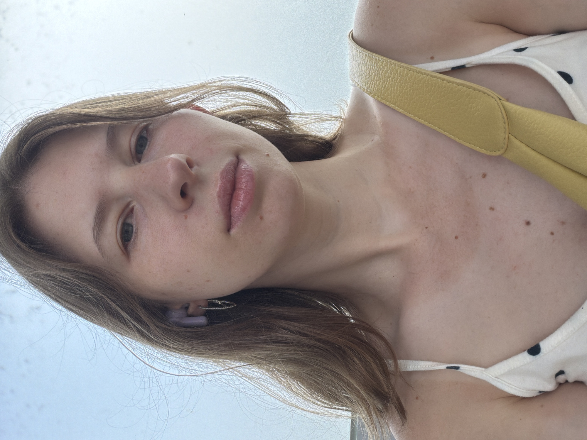 A young woman with shoulder-length brown hair and light skin is looking into the camera. She is wearing a white camisole with black polka dots and a yellow purse strap is visible across her right shoulder. She has on a silver hoop earring and a light purple earbud. The background is a bright, overcast sky. The image was taken outdoors during the day.