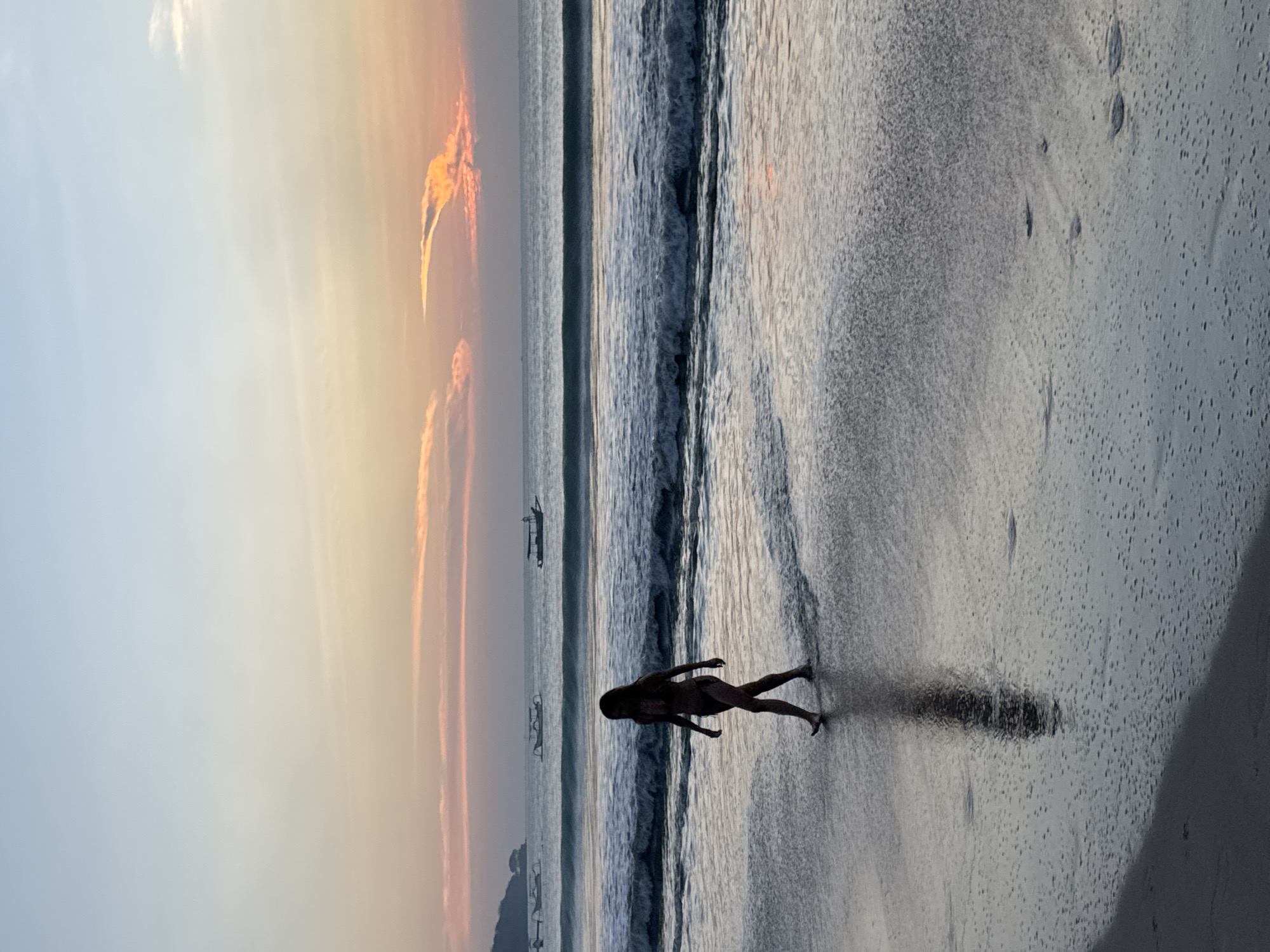 A woman in a bikini walks along the wet sand of a beach in Kuta Selatan, Indonesia. Her silhouette is dark against the muted colors of the sunset sky and the ocean. The sky is a gradient of pale blue at the top, transitioning to soft oranges and pinks near the horizon, with scattered, wispy clouds. The ocean has moderate waves breaking on the shore, and several small boats are visible in the distance. The wet sand reflects the sky and the woman's shadow, creating a shimmering surface. The scene captures a moment of solitude and natural beauty during twilight.