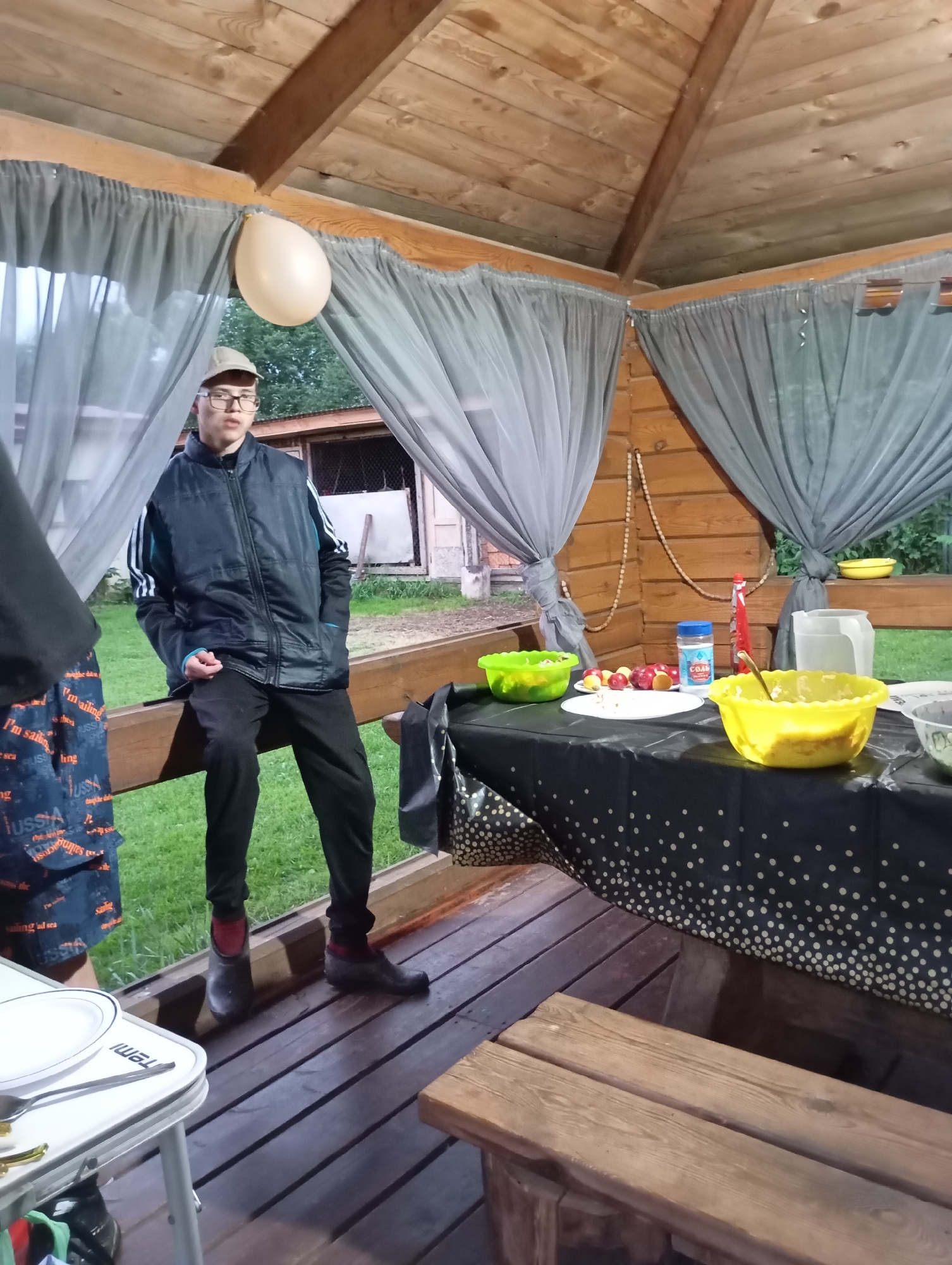 The image depicts an outdoor gathering under a wooden gazebo in Malyi Bukor, Russia. A young person wearing glasses and a baseball cap is seated on a wooden railing, dressed in a black puffer vest over a jacket with white stripes on the sleeves, black pants, and dark boots with red socks. To the left, another person's legs are visible, wearing patterned shorts. A table covered with a black tablecloth adorned with white polka dots is set up within the gazebo. On the table, there are two yellow bowls, one containing a green salad and the other a light-colored food, a plate with fruit, a jar with a blue lid labeled "СОЛЬ" (salt), and a red squeeze bottle, possibly ketchup. A large white jug is also present on the table. A white balloon is tied to the gazebo's ceiling. The environment appears to be grassy, with trees visible in the background. The overall scene suggests a casual outdoor meal or party.
