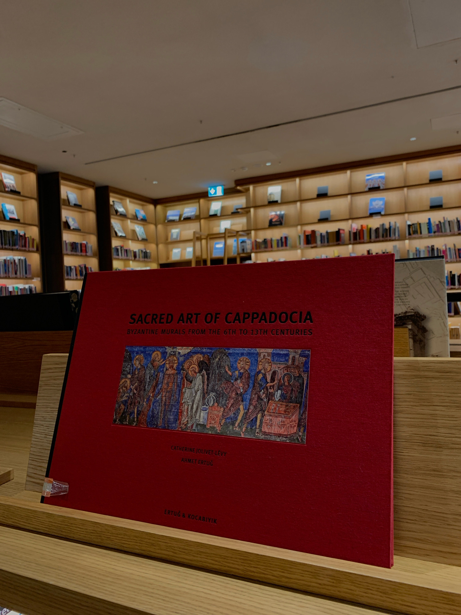The image shows a book titled "SACRED ART OF CAPPADOCIA BYZANTINE MURALS FROM THE 6TH TO 13TH CENTURIES" displayed on a wooden shelf in what appears to be a library or bookstore. The book has a bright red cover and features a detailed illustration of Byzantine artwork. The shelves in the background are filled with books, and a sign with a green arrow and a symbol of a person walking is visible on one of the upper shelves, indicating an exit. The setting suggests a modern, well-lit indoor space, possibly a cultural institution or retail establishment. The location is specified as Beyoğlu, Turkey.