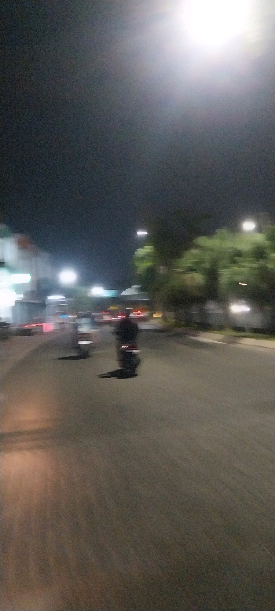 The image depicts a street scene at night in the city of Medan, Indonesia, characterized by motion blur due to movement during capture. In the foreground, a dark asphalt road is visible, with streaks of light suggesting reflected illumination. Two motorcycles are in motion on the road. The motorcycle on the right is more clearly defined, showing a rider with a dark top and dark pants, casting a long shadow on the road. The motorcycle on the left is less distinct, also appearing to be in motion. In the background, buildings with illuminated windows and streetlights line the left side of the road, creating a blurred urban environment. Trees with blurred foliage are visible on the right, and a bright, out-of-focus light source dominates the upper right portion of the image.