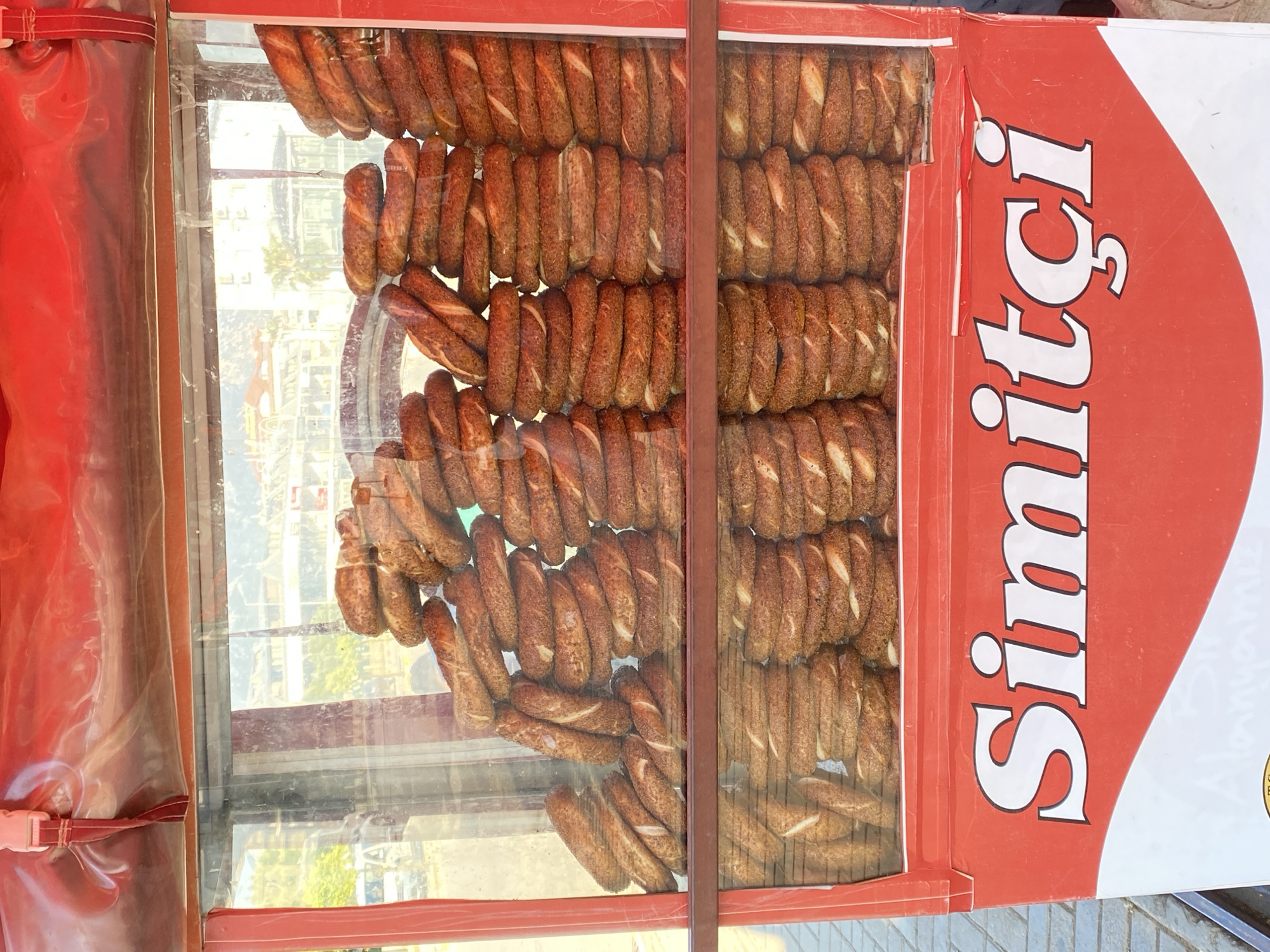 The image captures a vibrant display of simit, traditional Turkish bagels, arranged within a vendor's glass or plastic showcase. Hundreds of golden-brown simit, uniformly twisted and coated in sesame seeds, fill the frame, organized in neat rows and piles.
The scene depicts an outdoor setting, likely a street or market in Alanya, Turkey, given the context. Through the transparent display case, reflections of buildings and distant trees, possibly palm trees, are visible, indicating a bright, sunny daytime environment.
On the right side of the image, a prominent red sign with white lettering boldly reads "Simitçi," which translates to "Simit maker/seller" or "bagel vendor" in Turkish. This text clearly identifies the product being sold. The sign features a large white teardrop or abstract shape against its red background.
To the left, a section of red, possibly canvas or thick plastic, forms part of the vendor's stall or cart, secured with a white buckle. No people are visible, but the presence of the well-stocked display and signage suggests an active sales point. The focus is entirely on the array of freshly baked simit, ready for purchase.