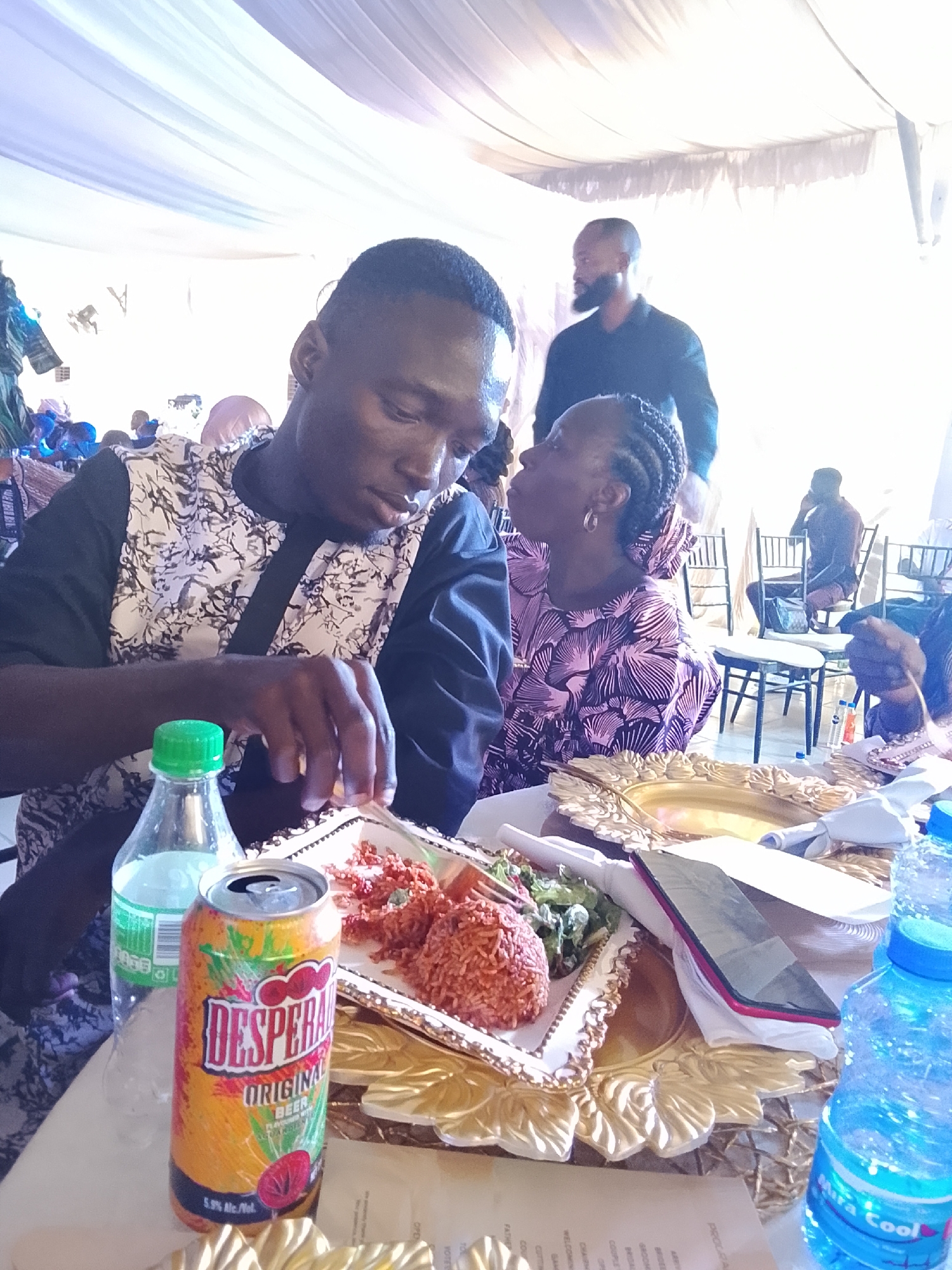 The image appears to be taken indoors at a social gathering in Abuja, Nigeria. A man in a black and white patterned shirt is seated at a table, appearing to be eating. He is looking down at a plate containing jollof rice, some other red sauce, and a small salad. To his right is a woman in a purple and black patterned dress, looking toward the man. On the table are several items including a can of Desperados beer, bottles of Mira Cool water, plates, a mobile phone, cutlery, and a menu card. In the background, other people are visible, including a man with a beard in a black shirt standing behind the seated woman. The setting is likely a wedding or party tent, indicated by the draped white material above. Chairs and tables with tablecloths are seen in the background.