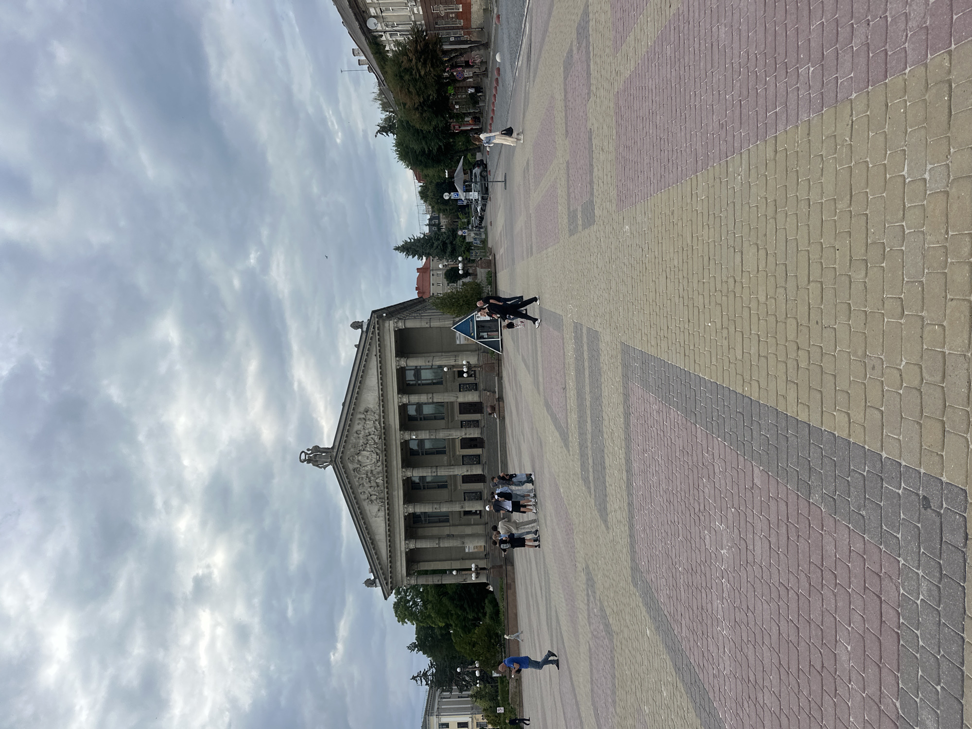 The image depicts the Ternopil Drama Theatre building in Ternopil, Ukraine, viewed from a paved plaza. The theater is a large, neoclassical structure with a prominent colonnade and a pediment adorned with sculptures. The plaza is paved with interlocking pavers in a geometric pattern of red, yellow, and grey. Several people are visible in the plaza, including a man walking in the foreground, a group of people standing near the theater, and a woman walking further in the distance. The sky is overcast with heavy clouds, and trees are visible on the sides of the plaza, lining the street and buildings in the background. Cars are parked along the street behind the plaza. The scene appears to be a public gathering space in front of a significant architectural landmark.