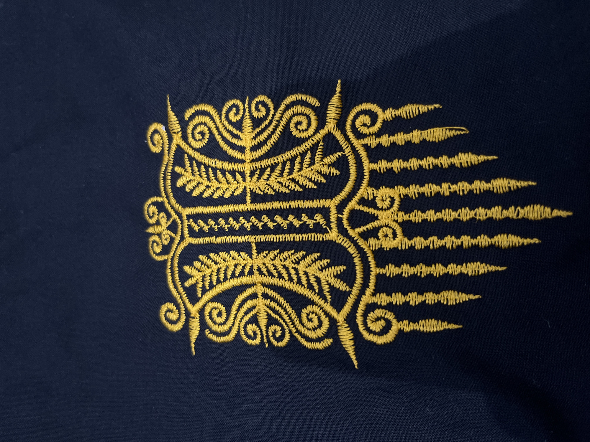 This media file presents a close-up view of intricate golden embroidery set against a dark blue fabric background. The embroidery, crafted with yellow-gold thread, features a detailed and symmetrical design. It includes prominent scrollwork, swirling patterns, and fern-like or stylized leafy motifs arranged horizontally.
On the right side of the main central design, a series of radiating, wavy lines extend outwards, giving the appearance of fringes or stylized rays. The visible stitching indicates skilled craftsmanship. The fabric appears to be a textile, possibly part of clothing or a decorative item.
There are no people, activities, or visible text in the image. The static nature of the shot provides no information regarding the time of day, weather, or emotional context. Given the location context of Lhokseumawe, Indonesia, the embroidery likely represents traditional or cultural patterns from that region.