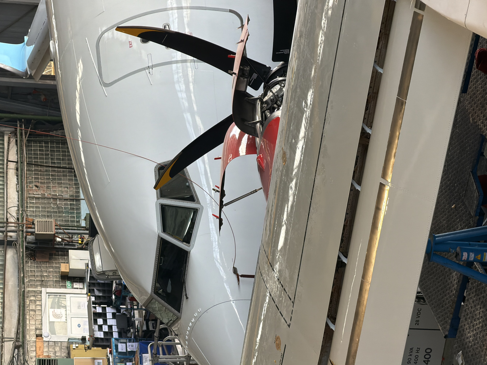 A wide-angle, slightly low-angle shot captures the front and right side of a white turboprop aircraft undergoing maintenance within a large hangar in Sofia, Bulgaria. The aircraft dominates the frame, showcasing its clean white fuselage, the cockpit windows, and a portion of the right wing. The engine nacelle on the right side of the fuselage is partially open, revealing internal engine components and a four-bladed propeller with yellow-tipped blades, indicating active maintenance or inspection work. Red accents are visible on the inner part of the engine cowling.
The background reveals a bustling industrial environment characteristic of an aircraft maintenance facility. To the left of the aircraft, various shelves filled with boxes and equipment are visible, alongside industrial wall structures made of glass blocks or metal grating. A ventilation unit is mounted on the wall. Underneath the right wing, a blue metal support structure is present. The lighting is artificial, suggesting an indoor setting.
Notable details include the crisp reflections on the aircraft's polished surface and the technical nature of the setting. Text visible on the aircraft includes "EXIT" in red lettering next to a door on the fuselage. On a component near the wing or under it, technical specifications are partially visible: "90 kVA", "400 Hz", and "28 VDC", along with a partial "400". No people are visible in the image.
