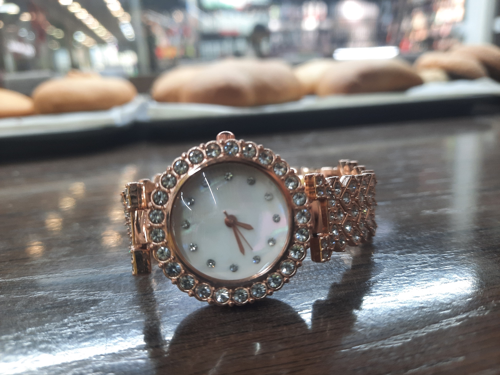 The image presents a close-up view of an elegant rose gold-toned women's wrist watch, positioned centrally on a dark, reflective surface. The watch features a circular face encircled by a bezel heavily adorned with numerous clear, sparkling rhinestones. The watch's face appears to be crafted from an iridescent material, possibly mother-of-pearl, and uses small clear rhinestones to mark the hours. The delicate hands, matching the rose gold tone of the watch, are visible, indicating the time is approximately 2:45. The watch band is intricately designed, also featuring multiple segments embellished with clear rhinestones, contributing to its luxurious aesthetic.
The surface beneath the watch is dark and highly polished, creating noticeable reflections of the watch and ambient light. In the softly blurred background, several light brown, rounded baked goods, resembling buns or pastries, are visible. These items are arranged on a tray or display rack, suggesting an indoor setting like a bakery, cafe, or a shop where such goods are sold. There are no individuals present, and no particular activity is depicted beyond the static arrangement of the watch. No visible text is present in the image, and the indoor lighting conditions do not provide clues about the time of day or external weather.