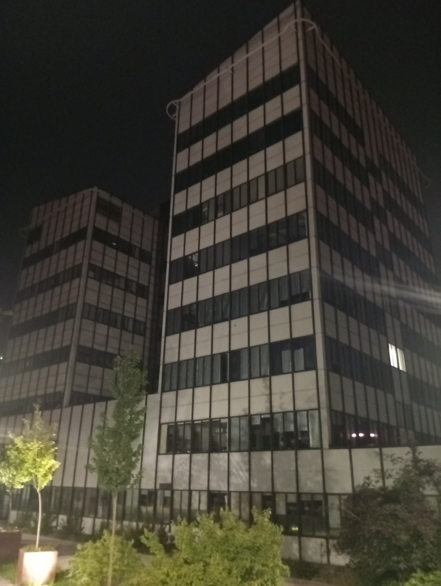 The image shows two modern office buildings at night in Wrocław, Poland. The buildings are tall, rectangular structures with a facade of alternating dark and light panels, creating a striped effect. They are illuminated by exterior lighting and some interior lights are visible through the windows, including a brightly lit window on the right-hand building. In the foreground, there are several trees and bushes, with some foliage illuminated by artificial light, suggesting a street or park setting adjacent to the buildings. The sky is dark, indicating nighttime. One of the buildings appears to have a decorative string of lights draped along its upper edge.