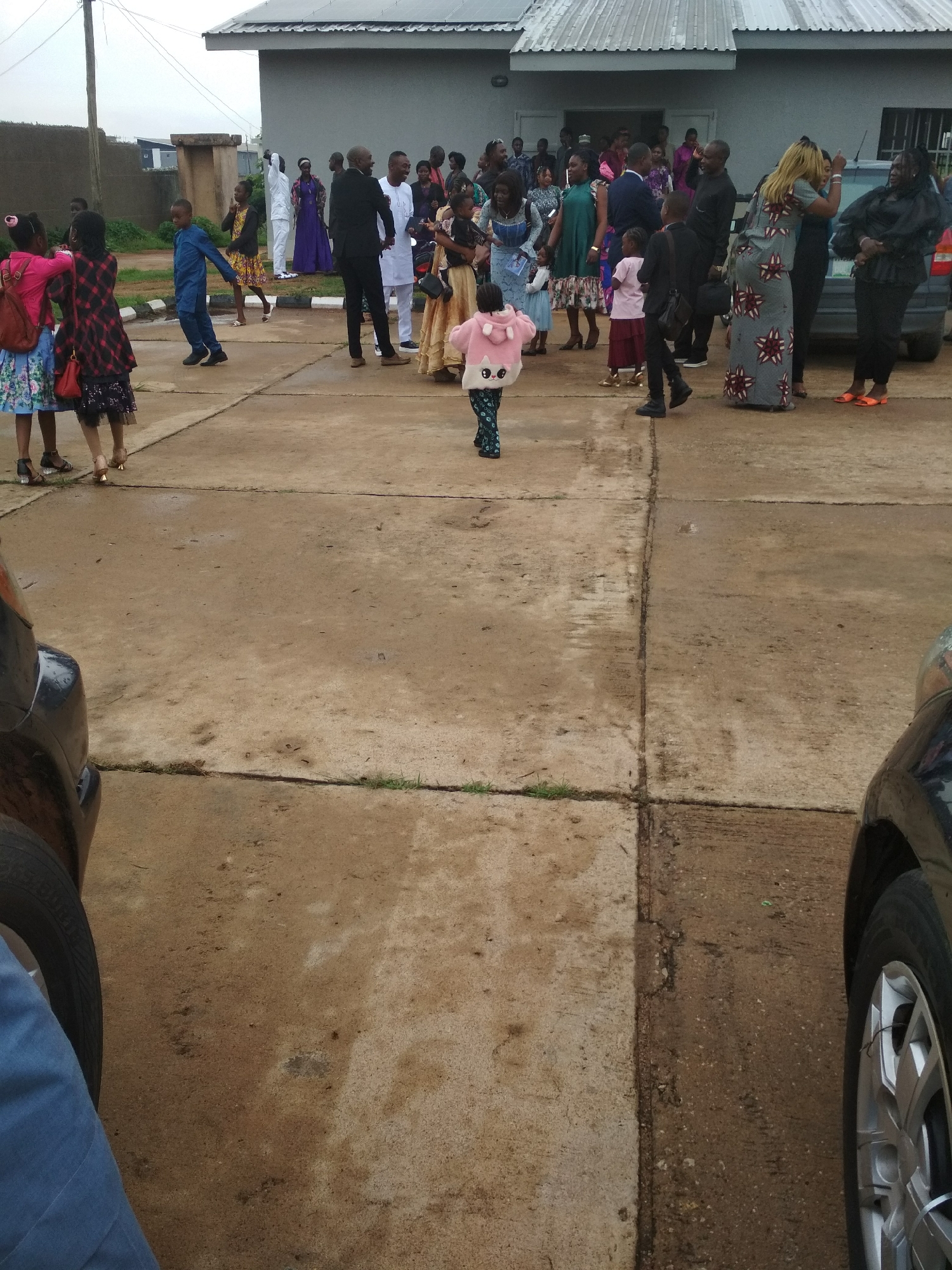 The scene depicts a gathering of people outside a building with a corrugated iron roof in Jos, Nigeria. The environment is a flat, paved area that appears to be wet. A group of individuals, dressed in a variety of formal and semi-formal attire, are congregated near the building's entrance. Several cars are visible parked nearby. In the foreground, a young child wearing a pink hooded jacket is walking away from the group. Other children are also present in the scene. Some people are interacting with each other while others are moving around.