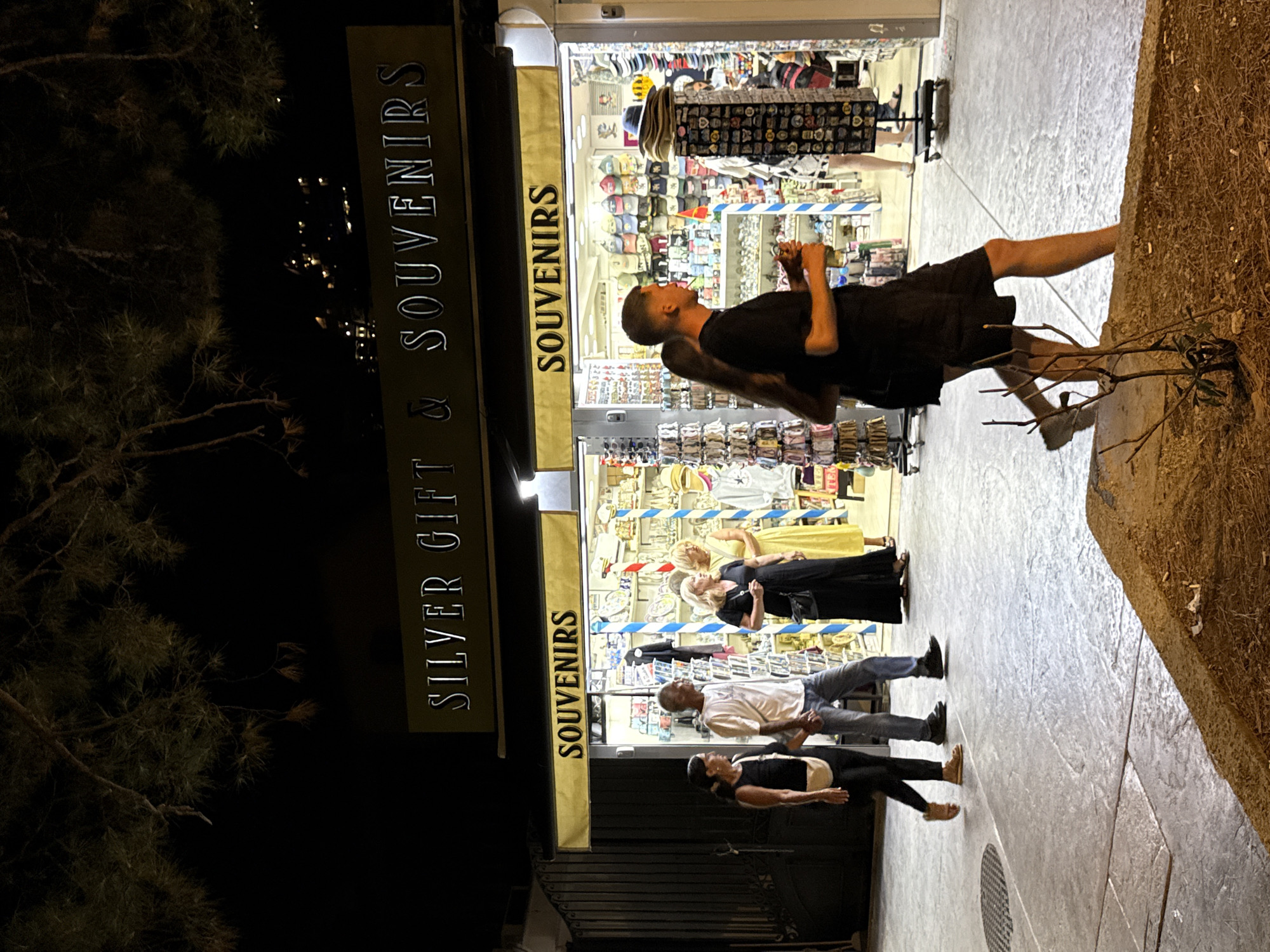 A nighttime street scene in Budva, Montenegro, shows a souvenir shop with bright interior lighting. A sign above the shop reads "SILVER GIFT & SOUVENIRS". Another sign, positioned below, says "SOUVENIRS" in larger lettering. Several people are walking past the shop. A couple in the foreground, a man and a woman, are walking away from the camera. The man is wearing a black t-shirt and shorts, and the woman is wearing a black dress. To their left, a couple is walking towards the camera. The woman is wearing a black tank top and leggings, and the man is wearing a white t-shirt and jeans. Further inside the shop, two women are visible. One is wearing a black dress, and the other is wearing a yellow dress. The shop displays a variety of merchandise, including hats, t-shirts, and trinkets. The street is paved with large, square concrete slabs. A small, dry bush is growing on a patch of dirt in the foreground. The overall atmosphere is that of a typical tourist street in a coastal town at night.