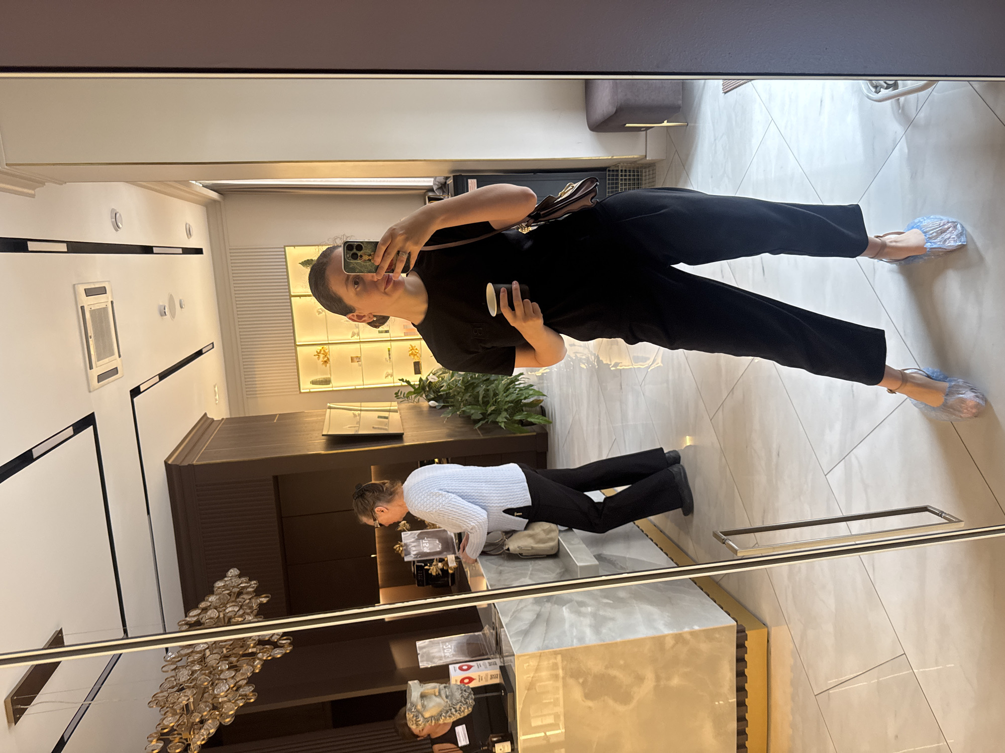 A woman stands in an indoor space, possibly a reception area or salon, taking a mirror selfie. She is wearing a black t-shirt, black trousers, and blue plastic shoe covers over high-heeled sandals. She holds a small cup in her right hand and her phone in her left, which is angled to capture her reflection.
Behind her, reflected in the mirror, is a reception desk with a marble countertop and a decorative bust on the left. To the right of the desk, a woman wearing a light blue sweater and black pants is standing, facing away from the camera and towards the desk.
The environment appears modern and well-lit, with a large mirror dominating the foreground. The ceiling features recessed lighting and what appears to be an air conditioning unit. In the background, there are shelves displaying products, a tall potted plant, and a dark brown wooden paneling on the walls. The floor is tiled with large, light-colored square tiles. The overall impression is of a clean, upscale commercial or service-oriented establishment.