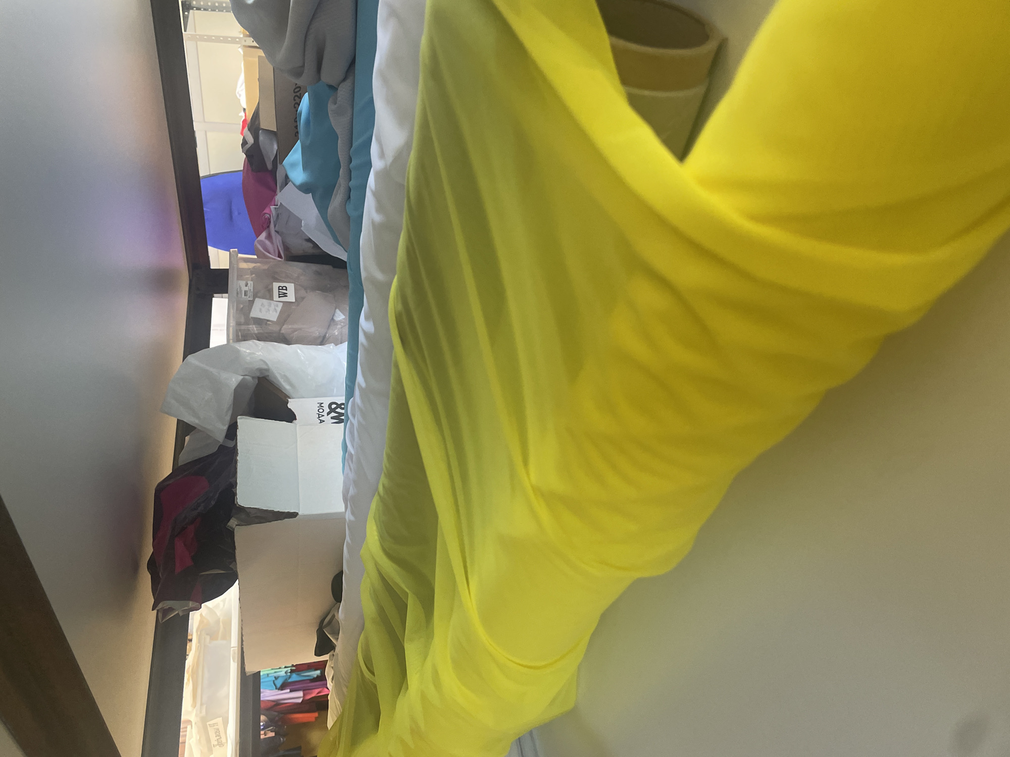 The image shows a storage area with shelves. On the shelves are various items including boxes, fabric, and clothing. In the foreground, there is a large roll of bright yellow fabric. In the background, there are shelves with boxes and folded fabrics in different colors. There are no people or actions depicted in the image. The location is unknown.