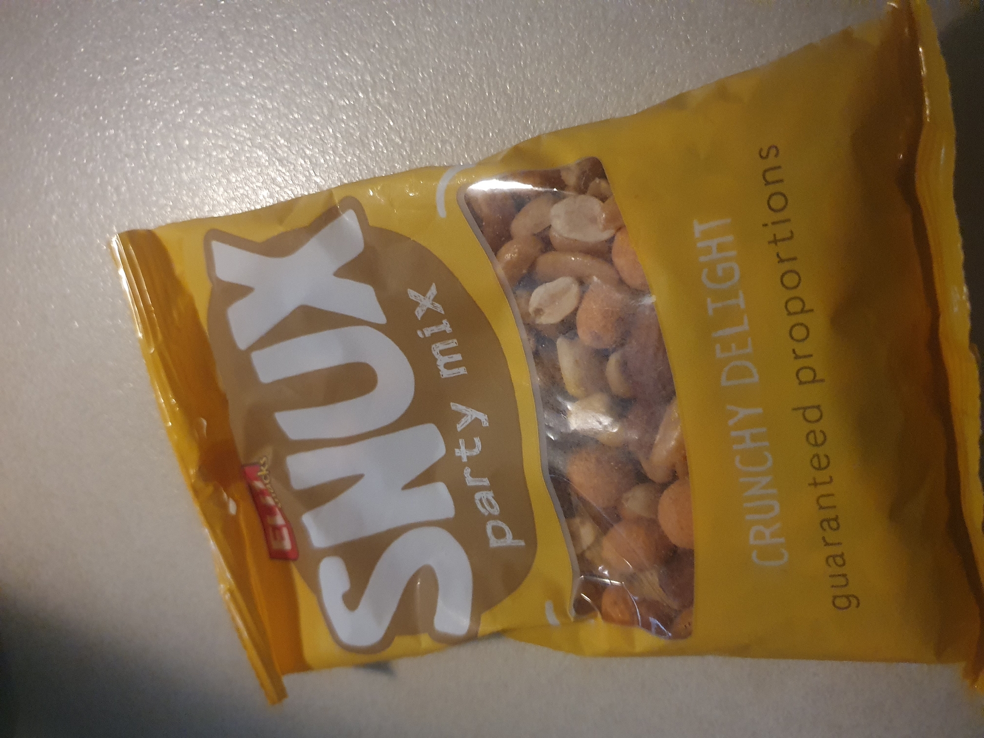 A partially open bag of "Sunks Party Mix" is lying on a light-colored, possibly metallic surface. The bag is yellow with a see-through section that reveals a mixture of nuts inside. The brand name "Sunks" is written in large, white, slanted letters on a tan background. Smaller text reads "party mix" and "CRUNCHY DELIGHT guaranteed proportions". A small red logo of "EVI Snacks" is also visible. The location is not identifiable as Plovdiv, Bulgaria.