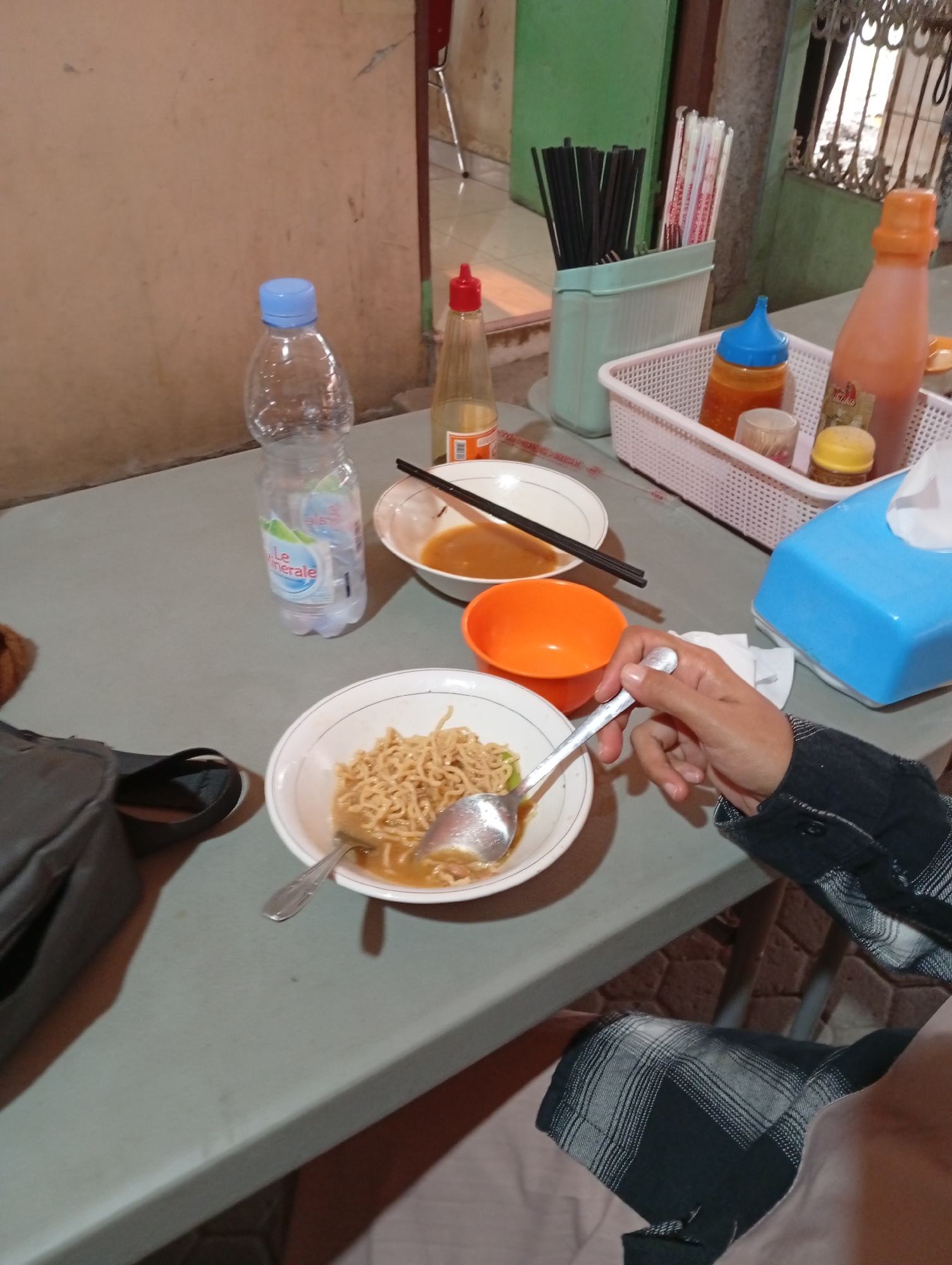 A high-angle, medium shot captures a table laden with food and condiments, likely in a food stall or restaurant in Curug Wetan, Indonesia. The scene is dominated by a gray tabletop.
In the foreground, a person wearing a black and white plaid shirt is visible, their right hand holding a spoon, about to pick up noodles from a white bowl. The bowl contains a portion of instant noodles in a savory broth, garnished with what appear to be green onions. To the left of this bowl is an empty orange plastic bowl.
Further up the table, a partially consumed bottle of "Le Minerale" water stands next to a white bowl containing a clear broth and chopsticks. Adjacent to this is a light blue tissue box, topped with a white tissue. Behind these items, a basket contains various condiment bottles, including a red-capped bottle of what seems to be chili sauce, a blue-capped bottle of sauce, and a small yellow-capped jar.
To the left, a dark gray bag is placed on the table. The background features a textured wall and a green cabinet. The overall impression is a casual dining setting.