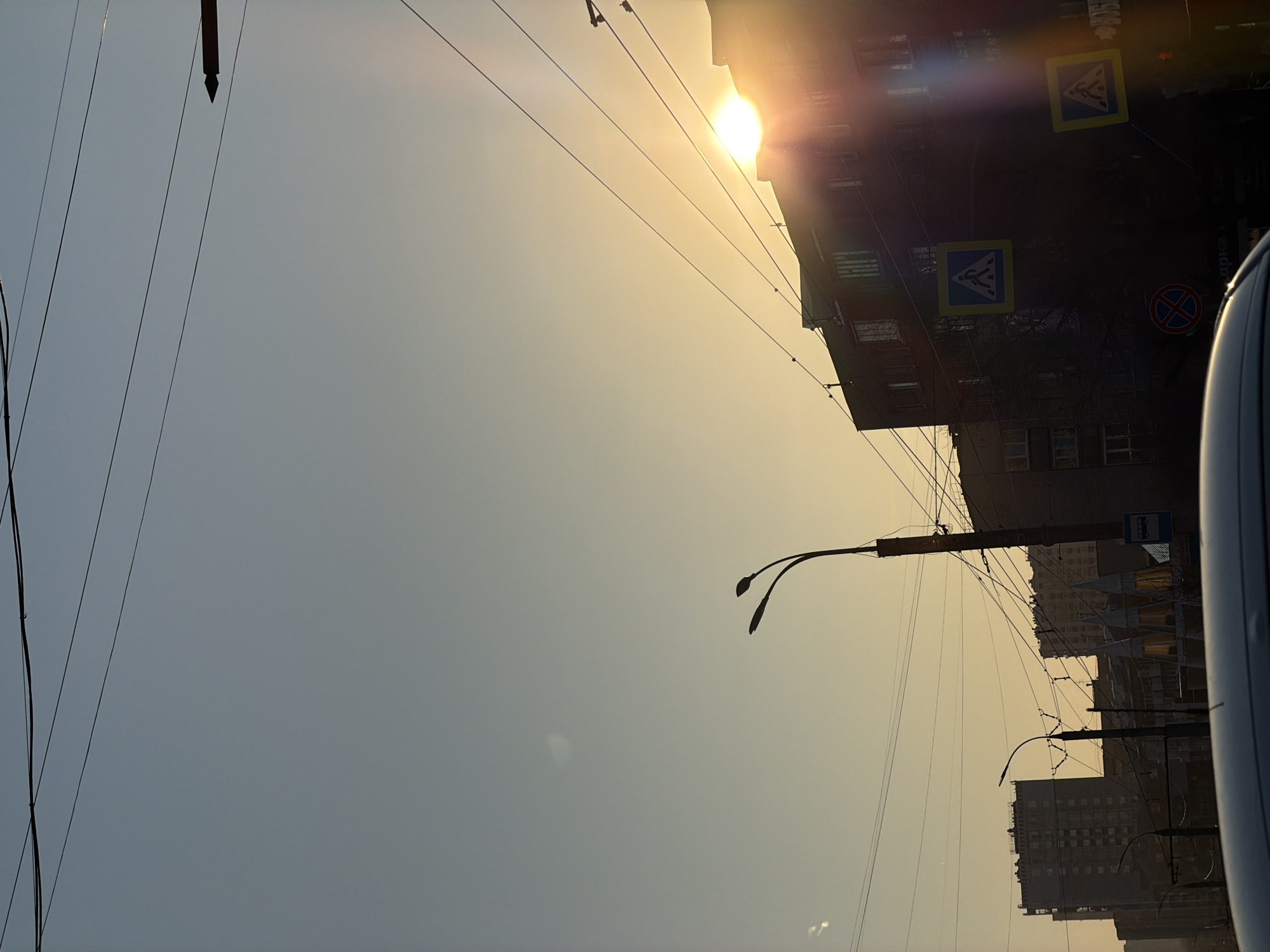 The image shows a street scene in Izhevsk, Russia, likely taken from inside a vehicle. The view is angled upwards towards the sky, which is a hazy yellow color, likely due to the sun. There is a building on the right side of the frame, partially silhouetted against the sun. Pedestrian crossing signs are visible on the building. Overhead power lines stretch across the sky. A street lamp is located in the middle of the frame. In the right side of the frame is part of a vehicle. Other buildings are visible in the distance.