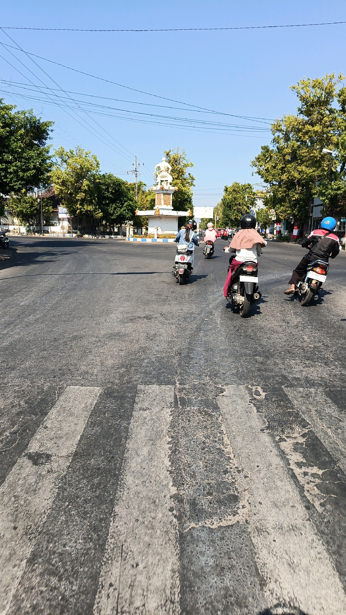 The image captures a street scene in Nologaten, Indonesia, on a clear, sunny day. In the foreground, a pedestrian crosswalk is visible, marked by worn, gray and white stripes on the asphalt road. Several motorcycles with riders are navigating the intersection.
In the middle ground, three motorcycles are moving away from the viewer. The first motorcycle, on the left, is white and ridden by a person wearing a blue denim jacket. Next is a dark-colored motorcycle carrying a rider with a light pink scarf over their shoulders. The third motorcycle, on the right, is black and ridden by someone wearing a black jacket and a blue helmet; this rider is looking back over their shoulder.
Further in the background, a white monument with a statue of a person on horseback stands in the center of the intersection, flanked by trees with green and yellow foliage. Buildings line the street on either side, and power lines stretch across the bright blue sky. The overall impression is of a typical day in a busy urban or semi-urban area of Nologaten.