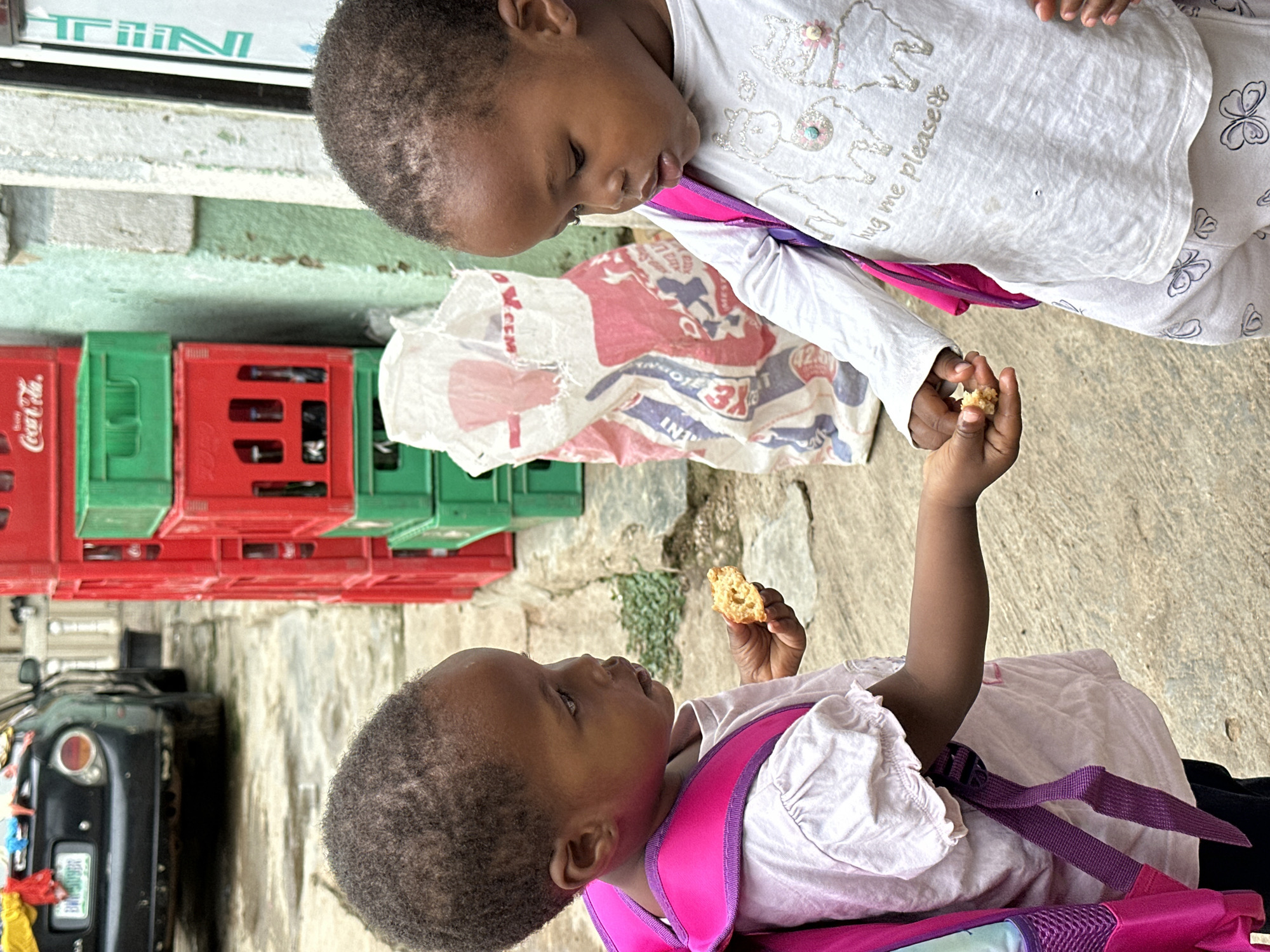 The image depicts two young Black girls interacting outdoors in what appears to be a commercial or residential area. The girl on the left, facing right, wears a light pink t-shirt with ruffled sleeves and a bright pink backpack. She is holding a small piece of food, possibly a biscuit or snack, in her right hand and offering it to the girl on the right. The girl on the right, facing left, wears a white long-sleeved shirt with llama graphics and the text "hug me please", and light-colored patterned pants. She is looking down at the food being offered.
Behind the girls, there are stacked red and green crates, some of which appear to be for beverages, with one displaying the "Coca-Cola" logo. A large white and red patterned bag is also visible leaning against a wall. In the background, a dark-colored car with a visible license plate is parked, and parts of a building with weathered walls and a white-framed window can be seen. The ground is paved with concrete. The setting suggests a street or an alleyway with elements of commerce. The overall atmosphere is candid, capturing a moment of sharing between the two children.