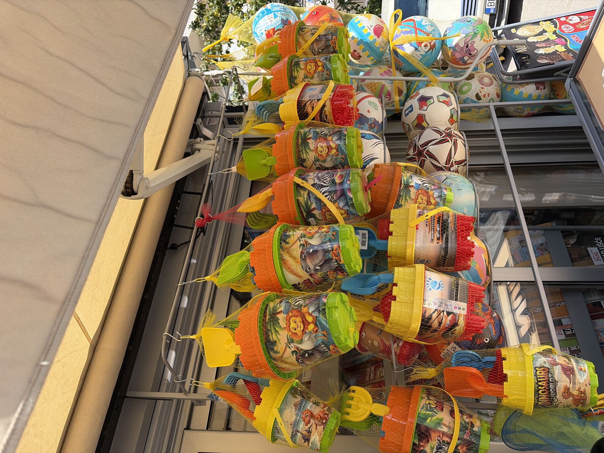 The image shows a display of children's beach toys, specifically buckets with shovels and rakes, along with various patterned balls, presumably for sale. The toys are hanging from a metal rack, suggesting they are located in a shop or outdoor vending area. A sign advertising ice cream and other frozen treats sits atop the display.
The scene appears to be outdoors, possibly near a tourist area, indicated by the presence of beach toys. The sign in the background indicates it is likely in the Balkans, specifically, in Budva, Montenegro. The sign includes street names and other local information. The image is taken from a low angle, looking up at the display.