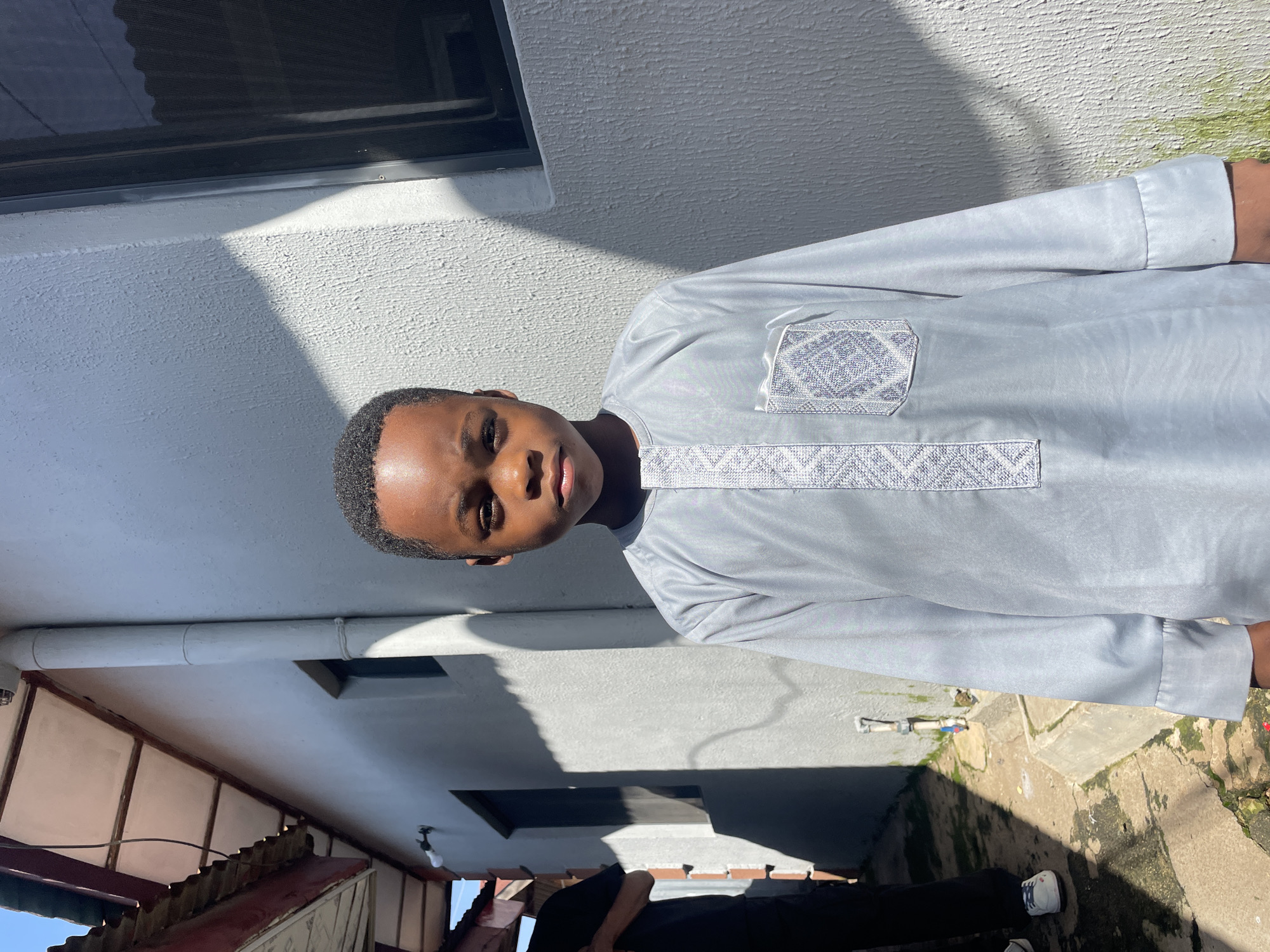 A medium shot shows a young boy in a light blue, traditional Islamic outfit, standing outdoors in what appears to be a narrow alleyway. The boy is positioned in the center of the frame, looking directly at the camera with a neutral expression. He has short, dark hair. The outfit features intricate white embroidery on the chest and a single breast pocket adorned with a patterned design.
The background consists of a textured, light gray wall on the right, with a dark-framed window. A shadow cast by an unseen object falls diagonally across the wall and the boy's clothing. To the left of the boy, a lighter colored building with a corrugated roof is visible, with a drainpipe running down the wall. Further back in the alley, the legs of another person wearing dark clothing and white sneakers can be seen. The ground in the alleyway is paved with uneven stones and shows signs of wear and tear, with patches of moss and dirt. The overall environment suggests an urban setting, possibly within a residential area in the city of Jos, Nigeria. The lighting indicates a bright, sunny day.