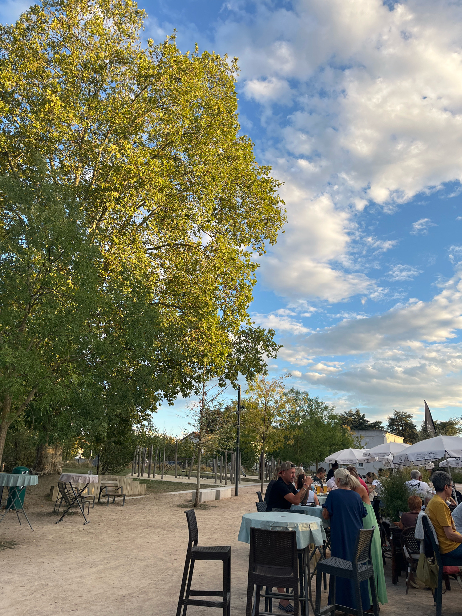 In Saint-Just-Saint-Rambert, France, an outdoor seating area is set up on a sandy ground, possibly a park or a communal space. Several tables with chairs are arranged, some occupied by people. To the left, a large tree with lush green leaves provides shade. In the background, a playground structure with wooden poles and ropes is visible. Further back, white umbrellas and more tables suggest a cafe or restaurant. The sky is blue with scattered white clouds, indicating fair weather.