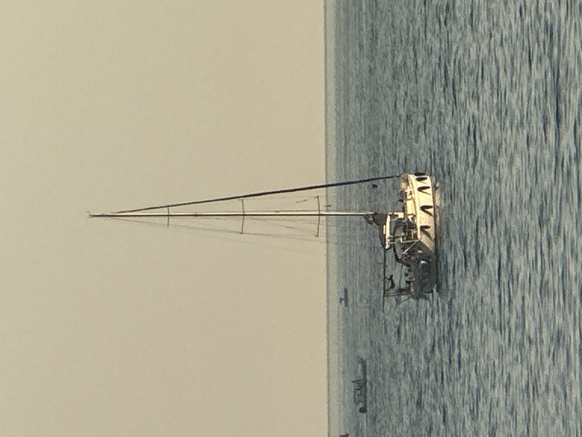 A sailboat with a tall mast and a white hull is anchored in calm blue water. Several other smaller boats are visible in the distance. The sky is a pale, hazy yellow, suggesting it is either early morning or late afternoon. The scene appears to be at sea.