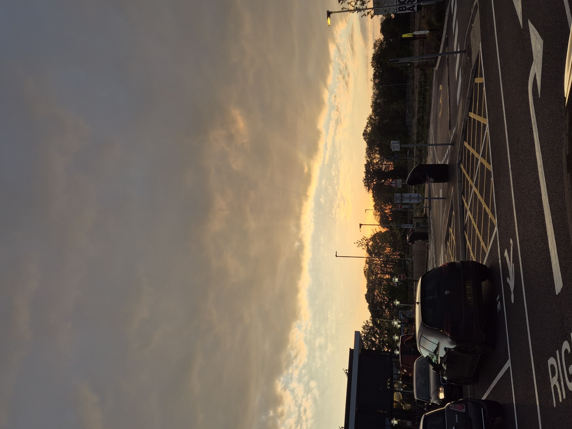 The image depicts a parking lot in West Suffolk, United Kingdom at dusk or dawn. The sky is a mix of dark clouds and golden light. The parking lot is paved and has painted parking spaces with directional arrows. Cars are parked in some of the spaces. Streetlights illuminate the area. Trees are visible along the edge of the parking lot and the background includes buildings and signage.