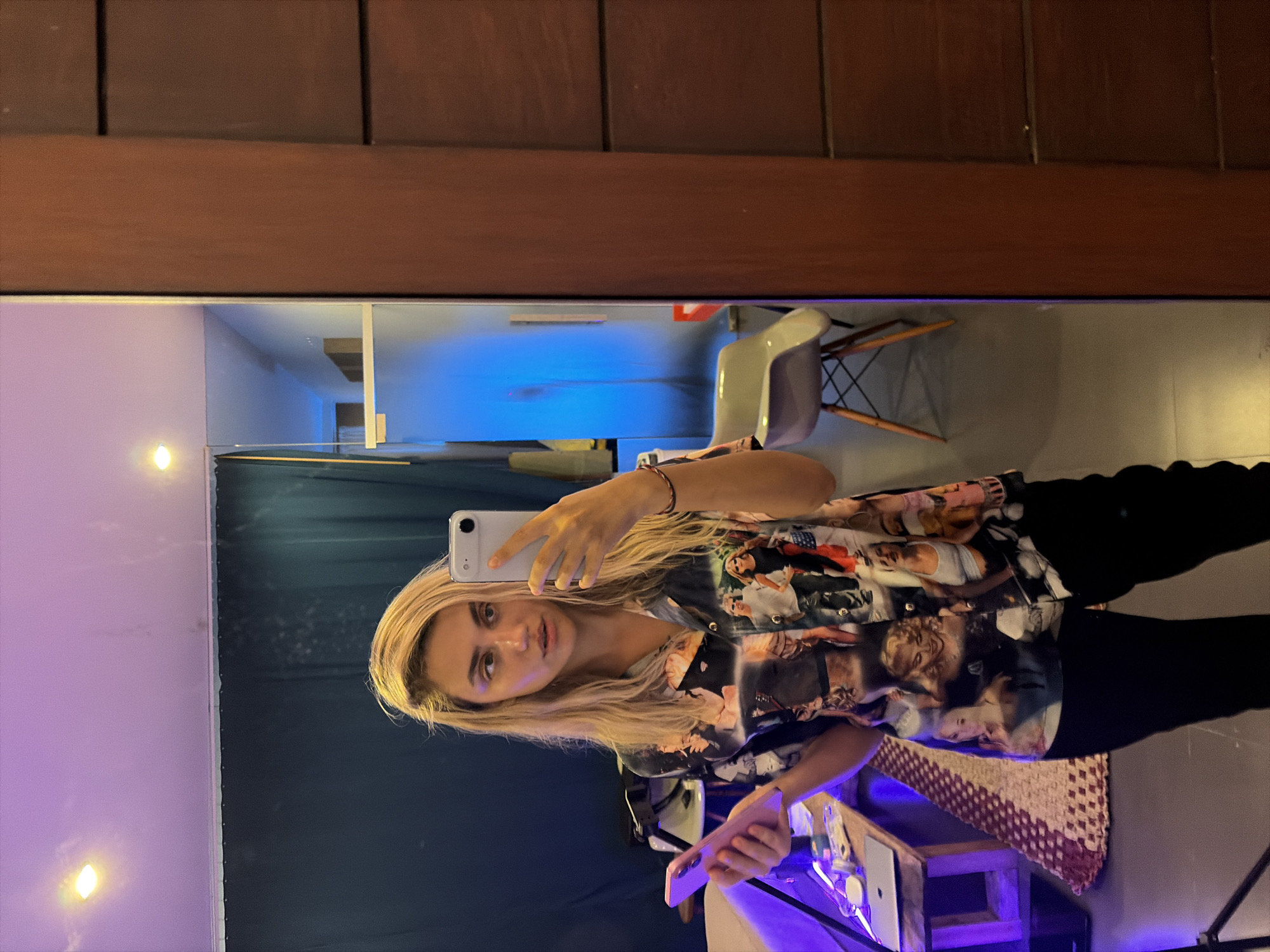 A young woman with blonde hair is taking a mirror selfie in what appears to be an indoor studio space. She is wearing a short-sleeved, collared shirt covered in a collage of images, predominantly of women, and black pants. In her right hand, she holds a silver smartphone, pointed towards the mirror. In her left hand, she holds a pink smartphone. The background shows a dark curtain, a wooden door frame, and a chair with a white seat and wooden legs. There is also a table with a laptop on it, and a rug. The lighting in the room includes purple and blue hues. The location is not specified in the image.