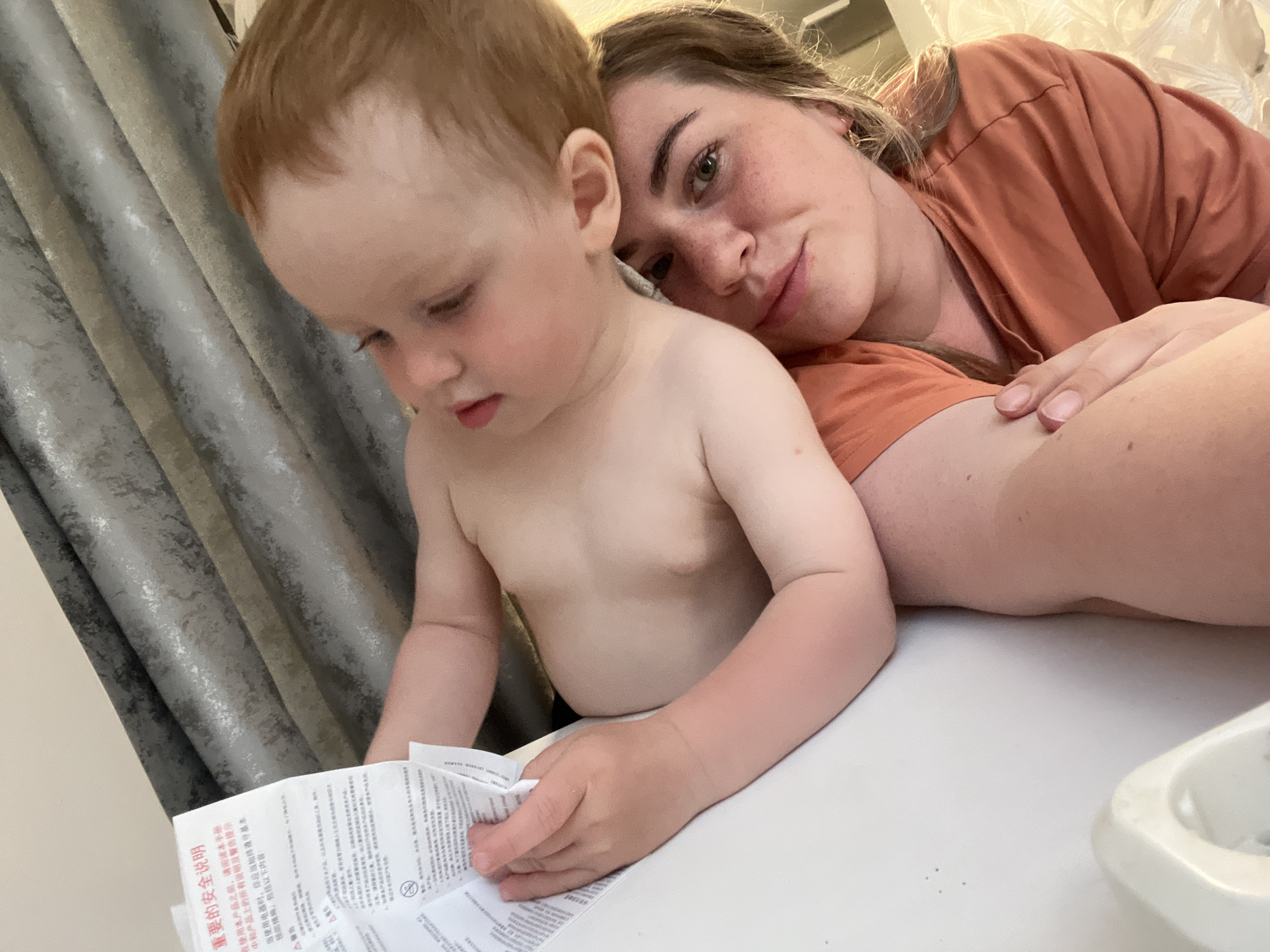 This is a close-up, indoor, medium shot of a woman and a young child on a white surface, possibly a table. The child, who appears to be a toddler with light brown hair, is shirtless and looking down at a piece of paper in their hands. The paper has text in what appears to be Chinese characters and some warning symbols. The woman, who has blonde hair and is wearing an orange shirt, is resting her head near the child, looking towards the camera with a gentle expression. Her arm is draped around the child. The background shows a grey, textured curtain. The lighting suggests it might be daytime. The scene evokes a feeling of intimacy and quiet interaction between the woman and child, possibly during a moment of reading or instruction. The visible text is in Chinese and appears to be safety instructions for a product. The location context is Chaikovsky, Russia.