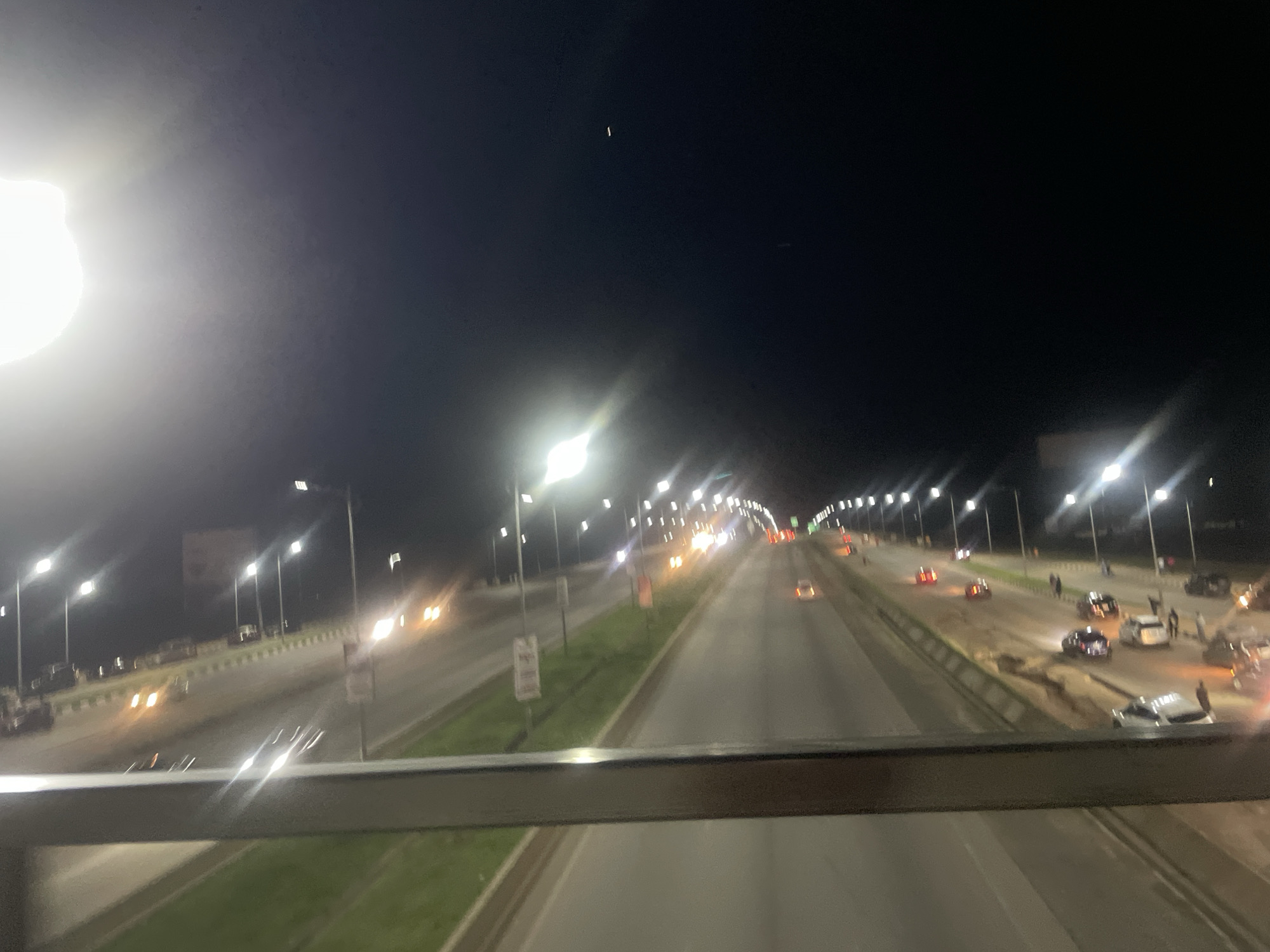 The image shows an aerial view of a highway at night in Sauka, Nigeria. The highway is illuminated by streetlights, and there are several cars on the road, some of which appear to be in a state of disrepair or have been involved in an incident. There are also people standing around some of the vehicles. The overall scene suggests a potentially chaotic or unusual situation on the highway.