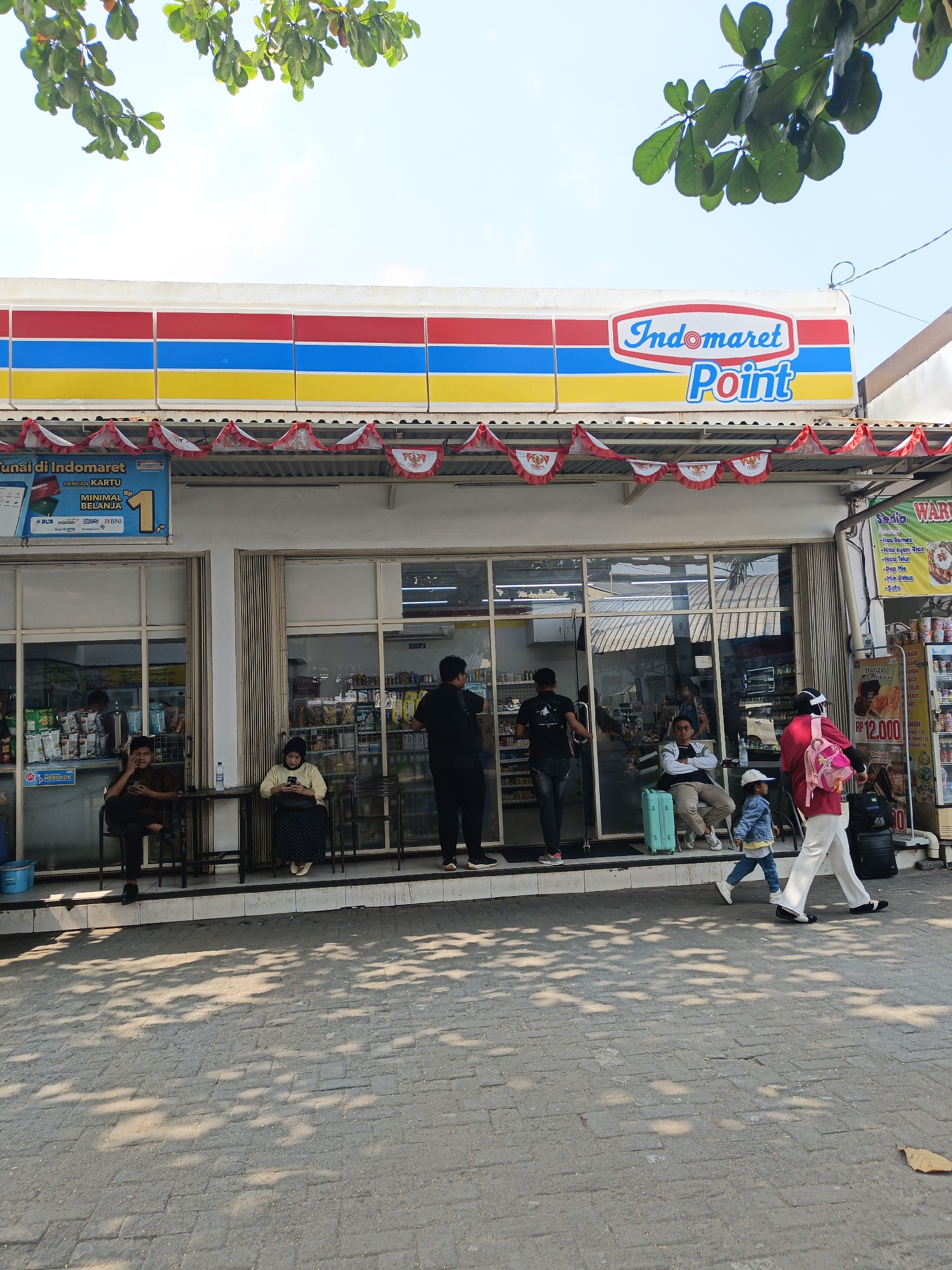 A frontal, eye-level, medium shot captures the storefront of an Indomaret Point convenience store in Surakarta, Indonesia, under a bright, clear sky with leafy branches from an overhanging tree casting dappled shadows on the paved sidewalk. The store's facade features a prominent red, blue, and yellow striped awning with the Indomaret Point logo in white and red text. A string of small Indonesian flag pennants decorates the top of the storefront.
Four individuals are visible outside the store. On the left, a man wearing a dark brown patterned shirt and black pants sits on a dark metal chair, looking at his phone. Next to him, a woman in a pale yellow long-sleeved shirt and a dark polka-dot skirt sits on another dark metal chair, also engaged with her phone. Further to the right, a man in a black t-shirt and black jeans stands looking into the store. To his right, a woman in a bright pink top, white pants, and a pink backpack walks past, her face partially obscured by a transparent face shield. She is accompanied by a young child in a denim jacket and blue jeans, walking in the same direction.
Inside the store, visible through large glass windows and sliding doors, shelves are stocked with various products. A light blue suitcase rests on the ground next to the man in the black t-shirt. A black suitcase is positioned near the walking woman.
The environment is a typical urban street scene with the store as the central focus. The sidewalk is paved with grey rectangular bricks. A sign with red and white text and an image of a food item is affixed to the wall to the right of the entrance. Another smaller sign with "DILARANG MEROKOK" (No Smoking) is visible on the left window.