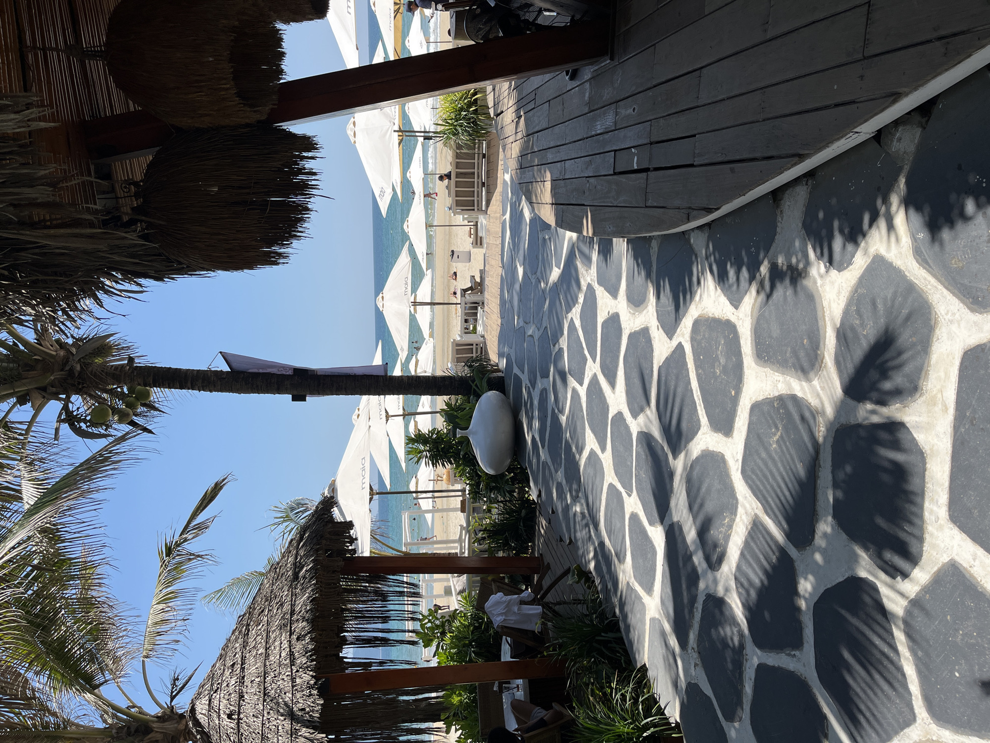 The image captures a view from an outdoor seating area in Đà Nẵng, Vietnam, possibly at a beachfront restaurant or resort. The perspective is from under a thatched roof structure, looking out towards a beach with white umbrellas and sun loungers. The foreground features a black and white patterned stone or tiled floor and wooden decking. A palm tree is visible on the left, and various plants are integrated into the seating area. The beach is populated with a few individuals, and the ocean meets a clear blue sky.