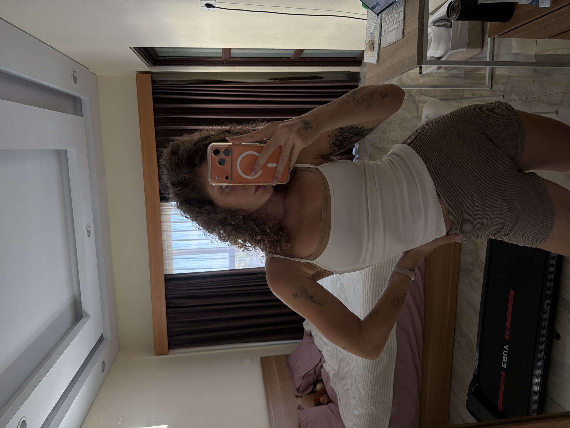 The image shows a woman standing in a bedroom, taking a selfie in a mirror. She is wearing a white tank top and brown ribbed shorts. Her hair is curly and brown. She has tattoos on her arms. In the background, there is a bed with purple pillows, a treadmill, a desk with a laptop, and a window with brown curtains. The room appears to be in a hotel or a rented apartment in North Kuta, Indonesia.