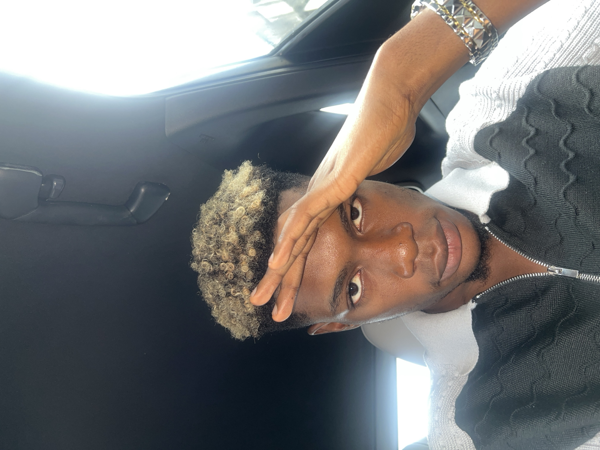 A close-up, slightly distorted, and rotated portrait of a Black man is presented. He is positioned inside a vehicle, indicated by the dark interior, including a door handle and a view of the sky through the car's roof. His curly hair is blonde-tipped. He is wearing a bracelet and a black and white textured jacket with a zipper. His right hand is raised, obscuring part of his forehead. The man is looking directly into the camera. The location is implied to be Lekki, Nigeria, but there is no visual evidence to support this claim. The image solely depicts the man within the interior of a vehicle.