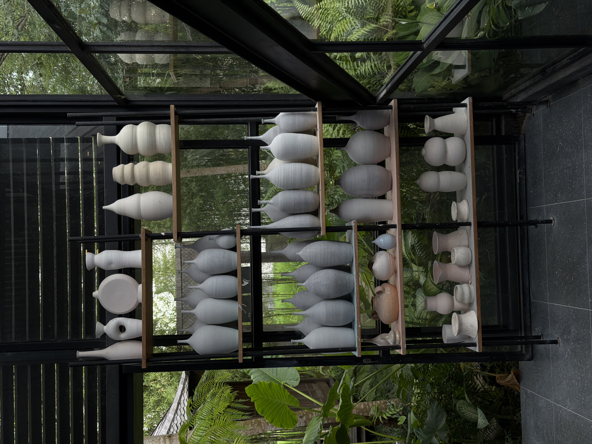 The scene depicts an interior view of a room, likely a display area or showroom, located in Singakerta, Indonesia. A tall, black metal shelving unit with five light-colored wooden shelves dominates the center of the image. The shelves hold an extensive collection of ceramic and pottery items, primarily vases and pots of various shapes and sizes. The pottery is predominantly unglazed, displaying tones of off-white, cream, and light grey, with some items exhibiting a speckled or textured finish. Shapes range from tall and slender to bulbous, conical, and abstract forms, including one vessel with a circular cutout. One terracotta-colored pot and one small blue-grey pot are visible on the third shelf from the top. The floor is covered with dark grey tiles or large paving stones. To the right, large windows with black frames provide a view of a lush, tropical exterior landscape featuring dense green foliage, large-leafed plants, ferns, and bamboo. A glimpse of a traditional dark-tiled roof is visible through the window in the upper left background. No individuals or active interactions are present in the scene.