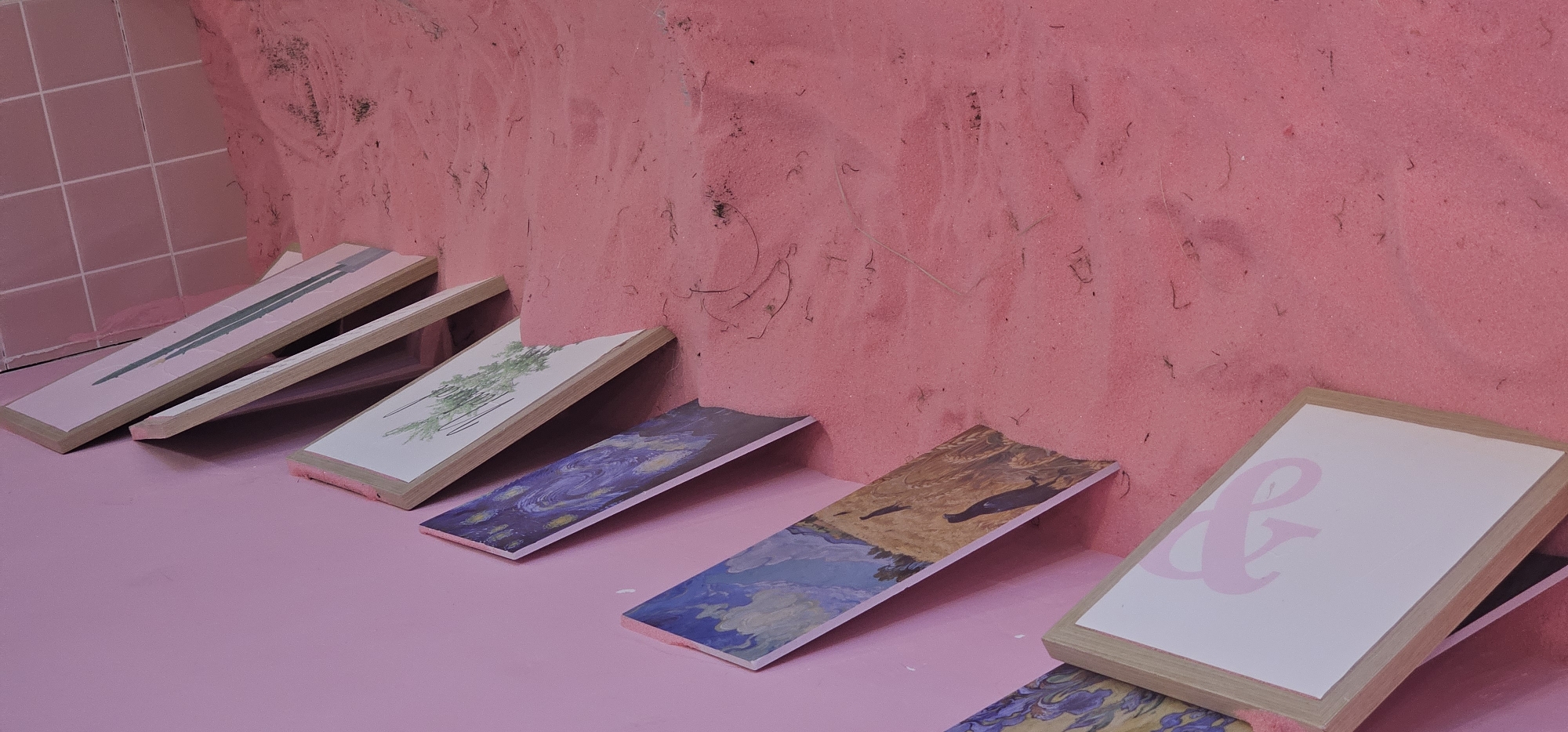 The image shows a collection of framed artwork displayed against a pink wall and resting on a pink sand-covered surface. The artwork appears to be leaning against the wall and partially embedded in the sand. The artwork includes what looks like a print of Van Gogh's "Starry Night," another print with a landscape featuring dark figures, and a minimalist print with green brushstrokes. The closest piece of artwork is a framed white surface with a pink graphic. The background consists of a tiled wall in a lighter shade of pink. The scene is indoors, and the pink sand suggests an artificial environment, possibly an art installation or display. There are no people visible in the image. The location is specified as Shapingba District, China, though this context is not visually apparent from the image itself.