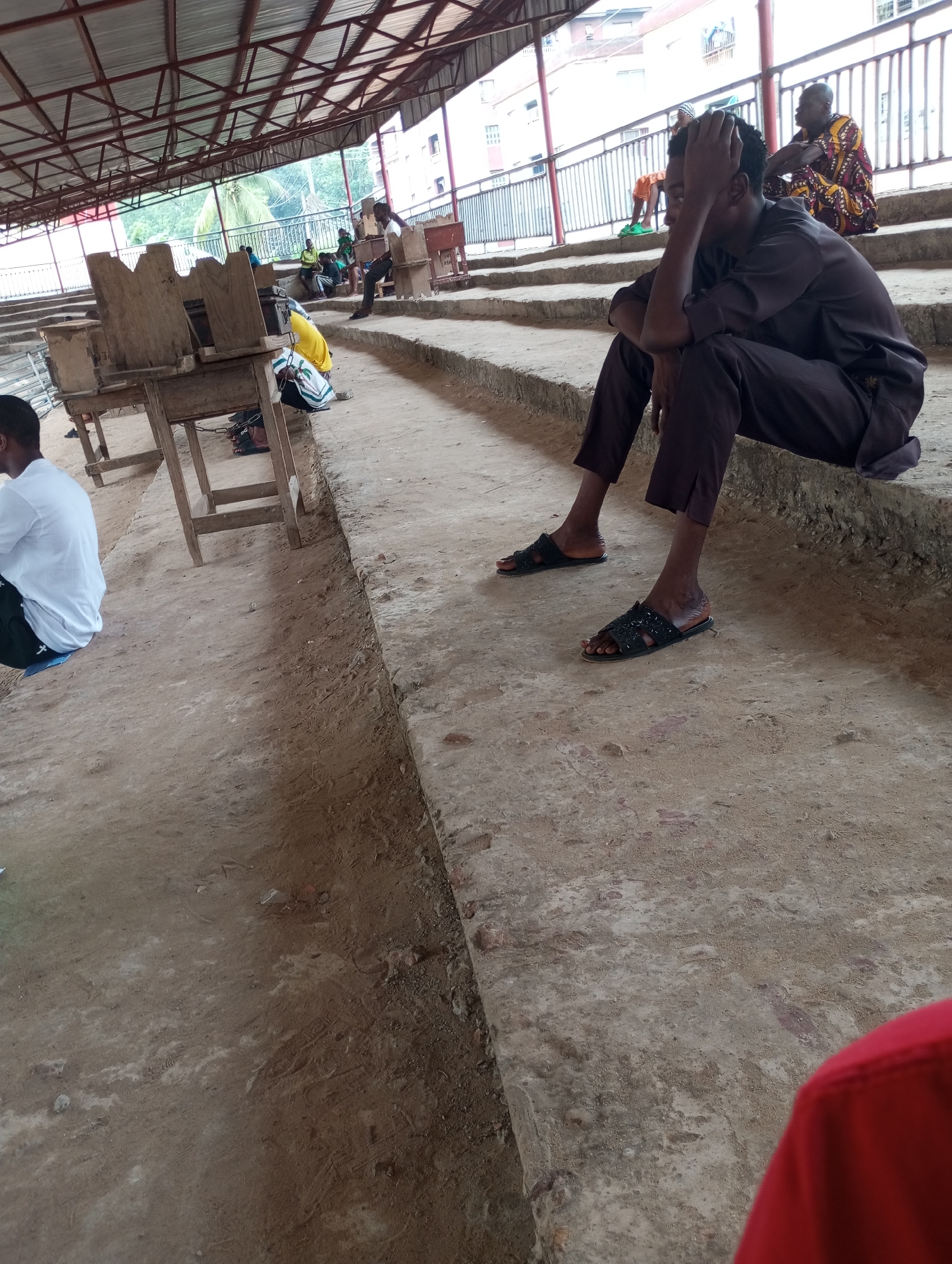 The image depicts a covered outdoor marketplace, likely in Onitsha, Nigeria. The roofing structure, made of corrugated metal and supported by metal beams, is visible overhead. The ground is a mixture of dirt and concrete. In the foreground, a young man in a dark brown outfit and black sandals sits on a concrete step, with his hand on his head. To his left, another individual in a white shirt and dark pants is seated on the ground, facing away from the camera. In the background, other people are seen sitting on steps, including a woman in a colorful patterned dress. Wooden stalls, some appearing to be empty and others with items on them, are also present. The environment suggests a busy but somewhat makeshift commercial area.